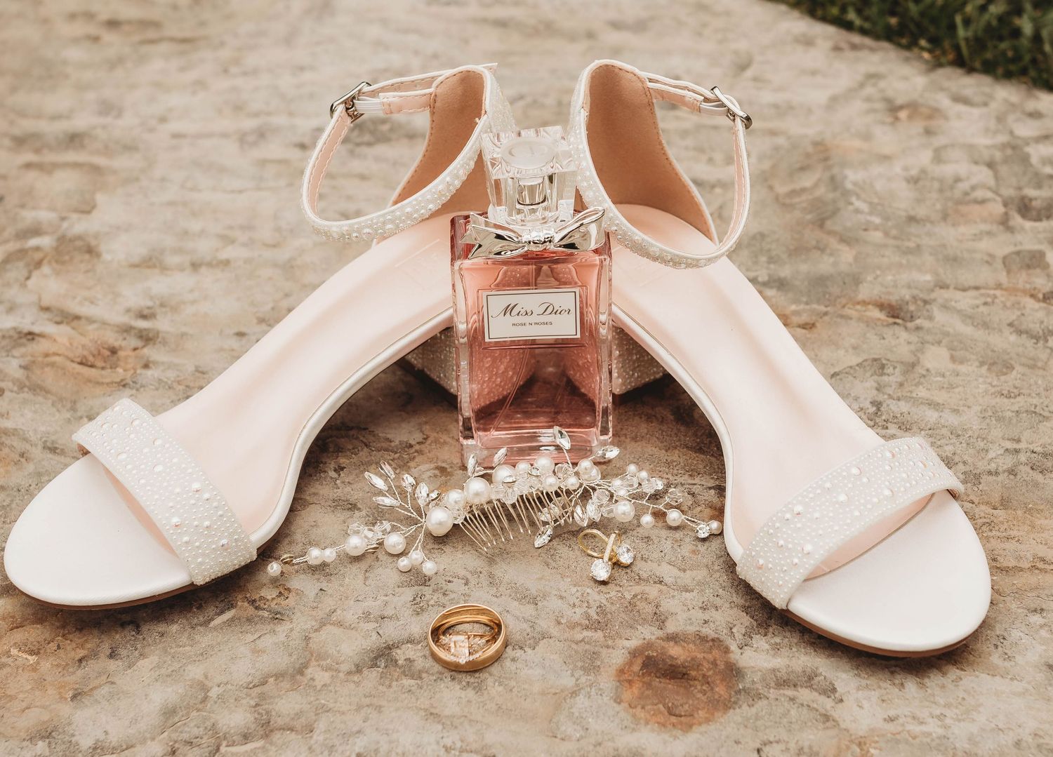 Wedding accessories including rings, sandals, and perfume arranged on textured stone.