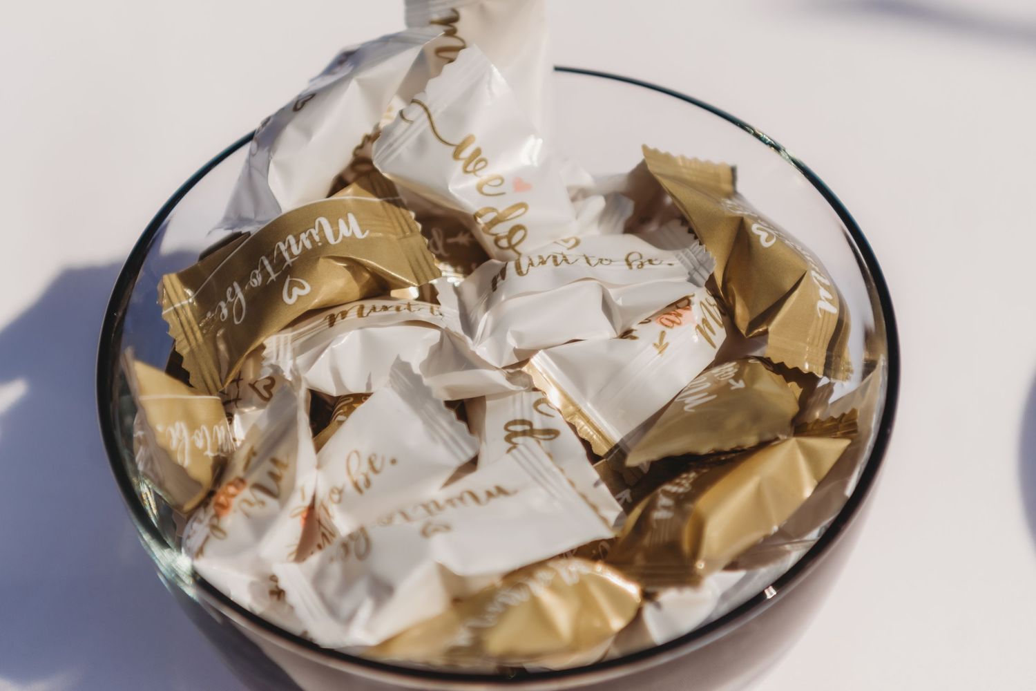 Glass bowl filled with gold and white wrapped chocolates candies on a white surface.