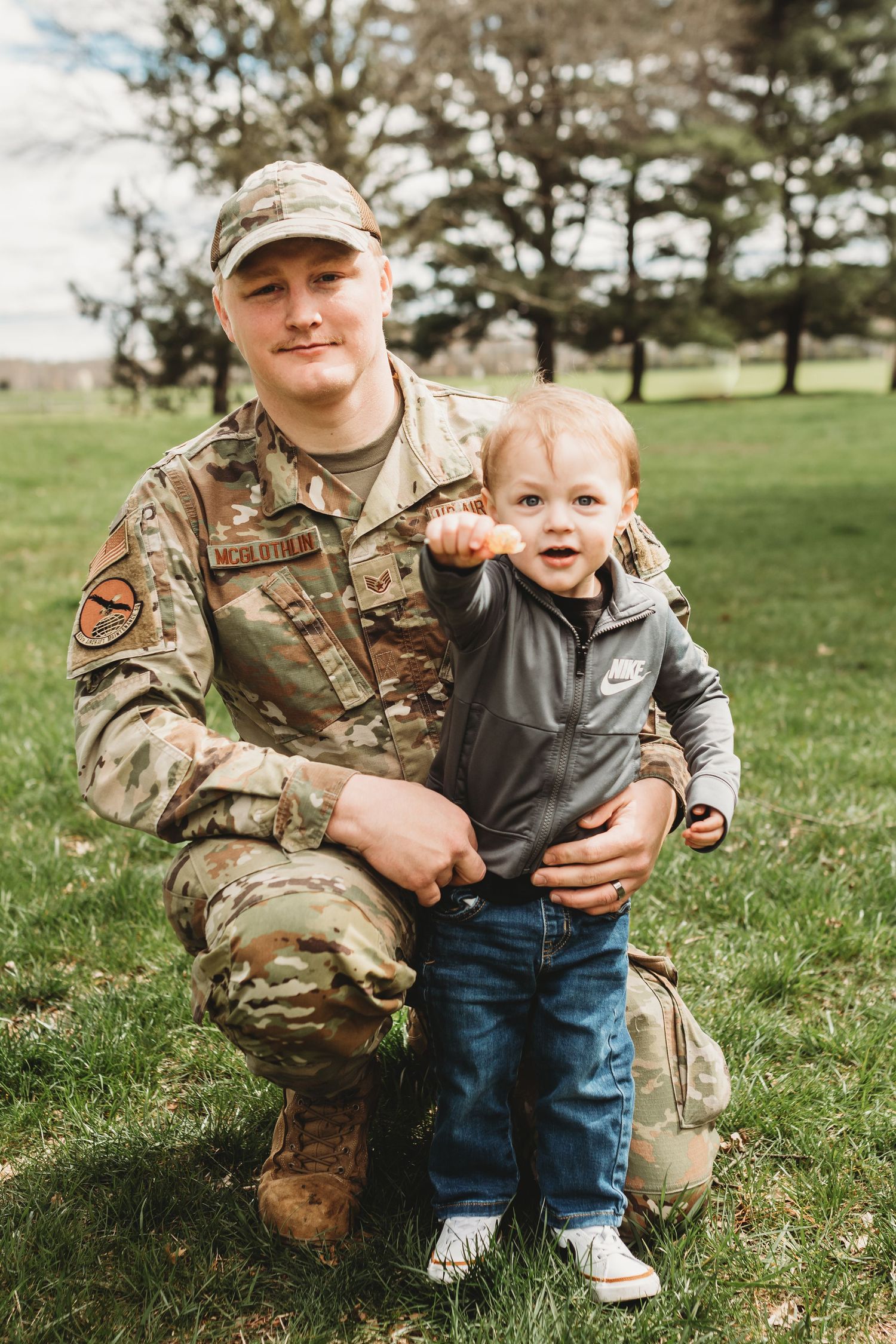 A service member in camouflage uniform kneels on grass beside a small child wearing jeans and sneakers.