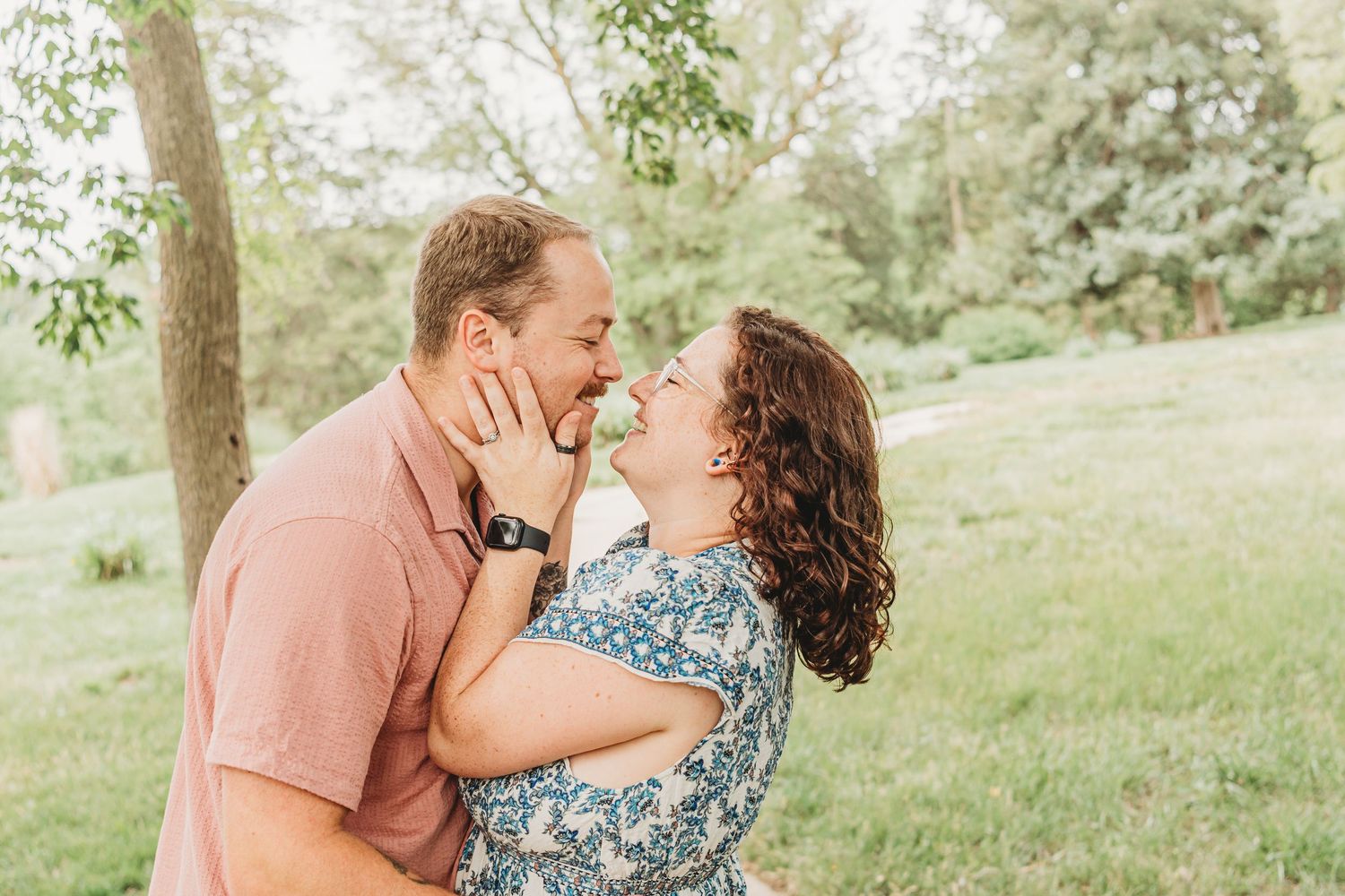 A couple shares a romantic moment outdoors, nearly kissing while embracing in a lush green park setting.