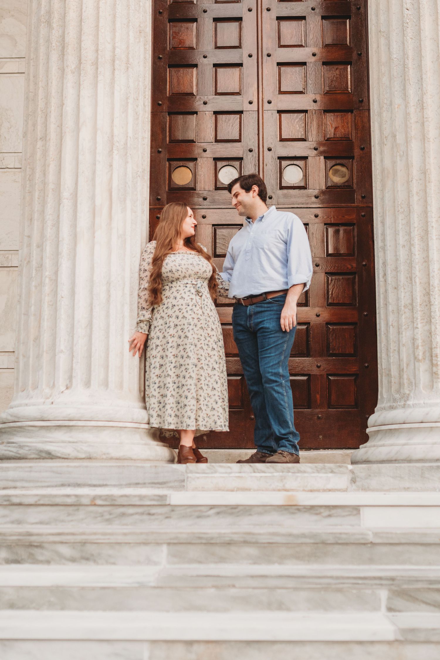 Couple poses for engagement photos in front of classic stone columns and wooden doors.