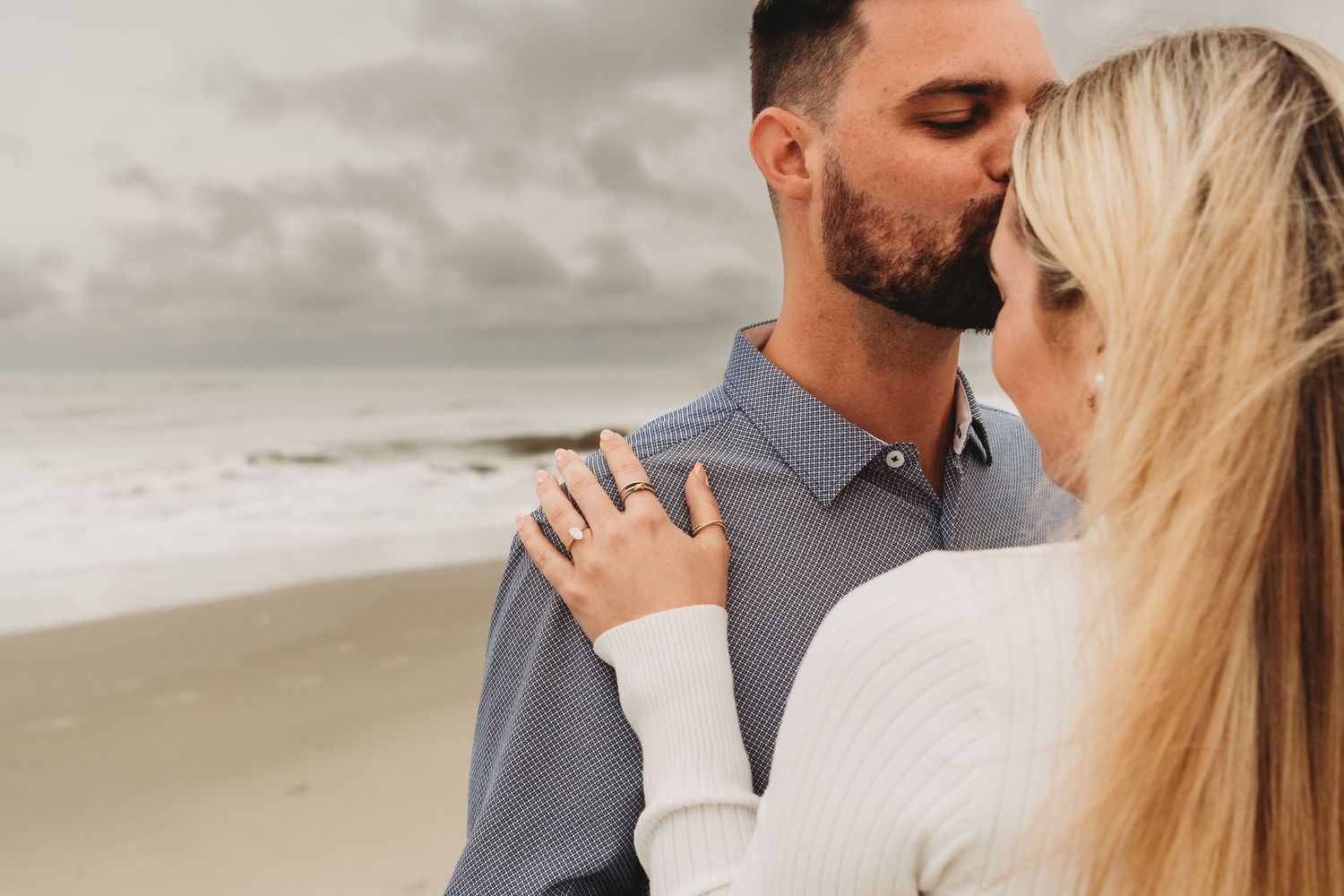 Couple sharing a romantic kiss on a beach with ocean waves and cloudy sky in the background.
