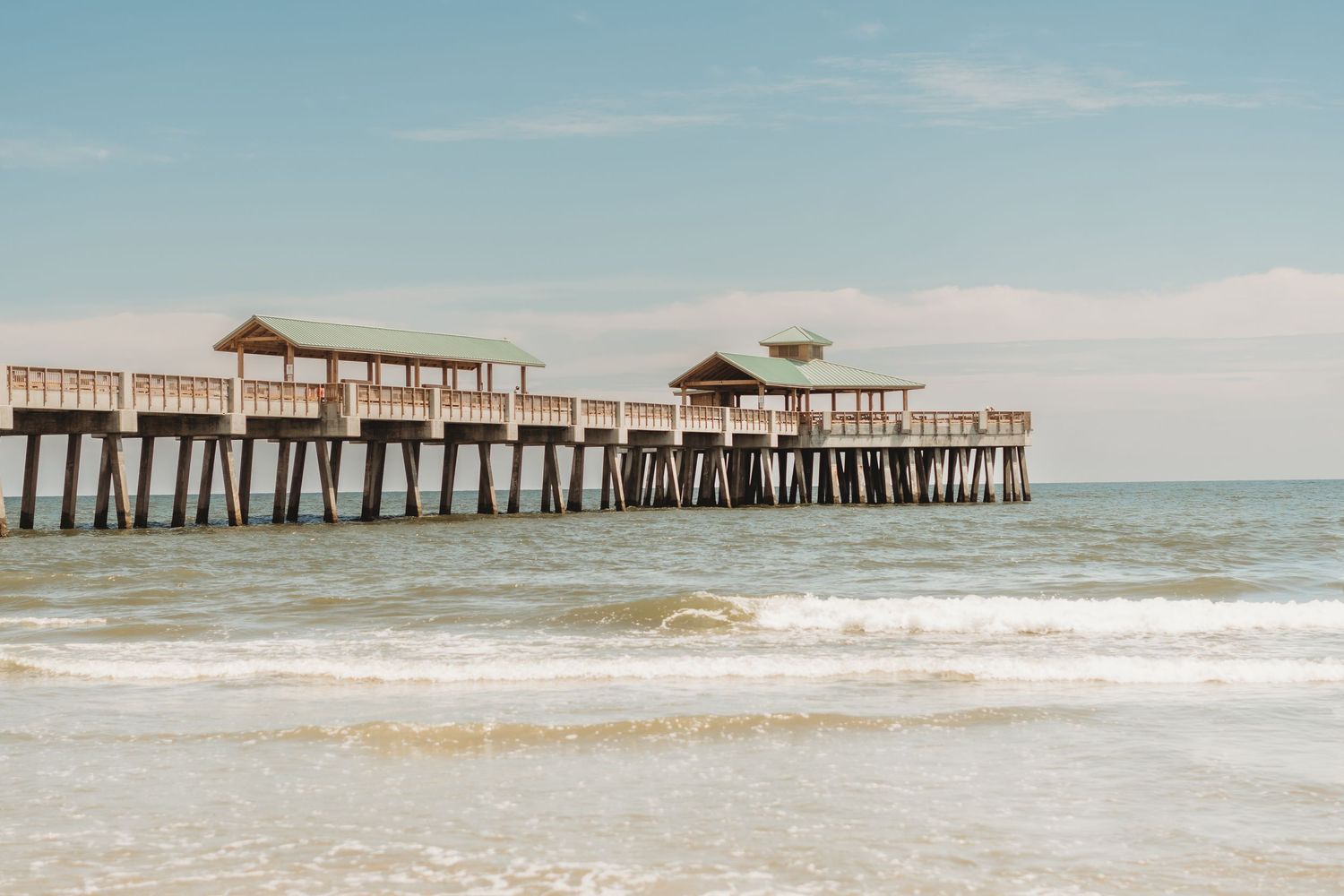 Wooden beach pier extending over calm ocean waves under a blue sky at a sunny coastal destination.