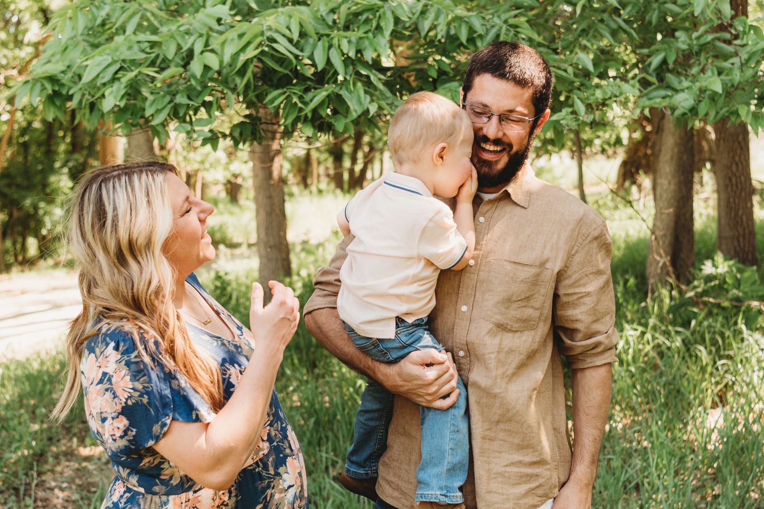 A happy family with a bearded dad holding a baby boy while a blonde mom laughs outdoors among green trees.