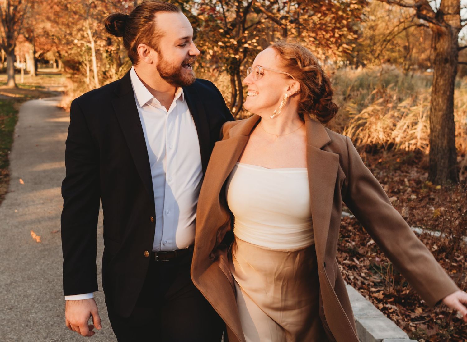 Couple smiling at each other while walking outdoors in fall, wearing elegant coats among autumn foliage.