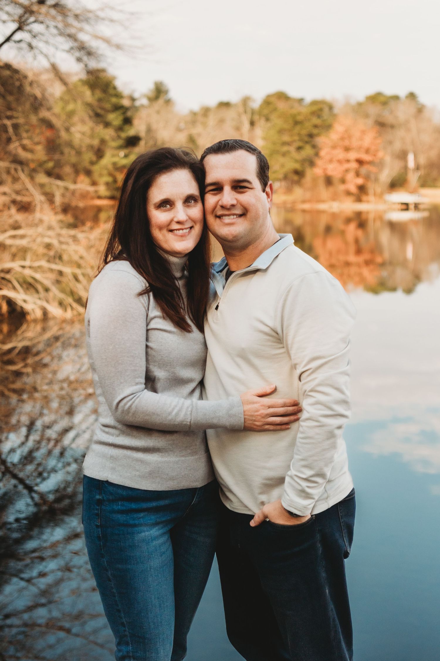 Couple in matching light sweaters laughing together by autumn lake.