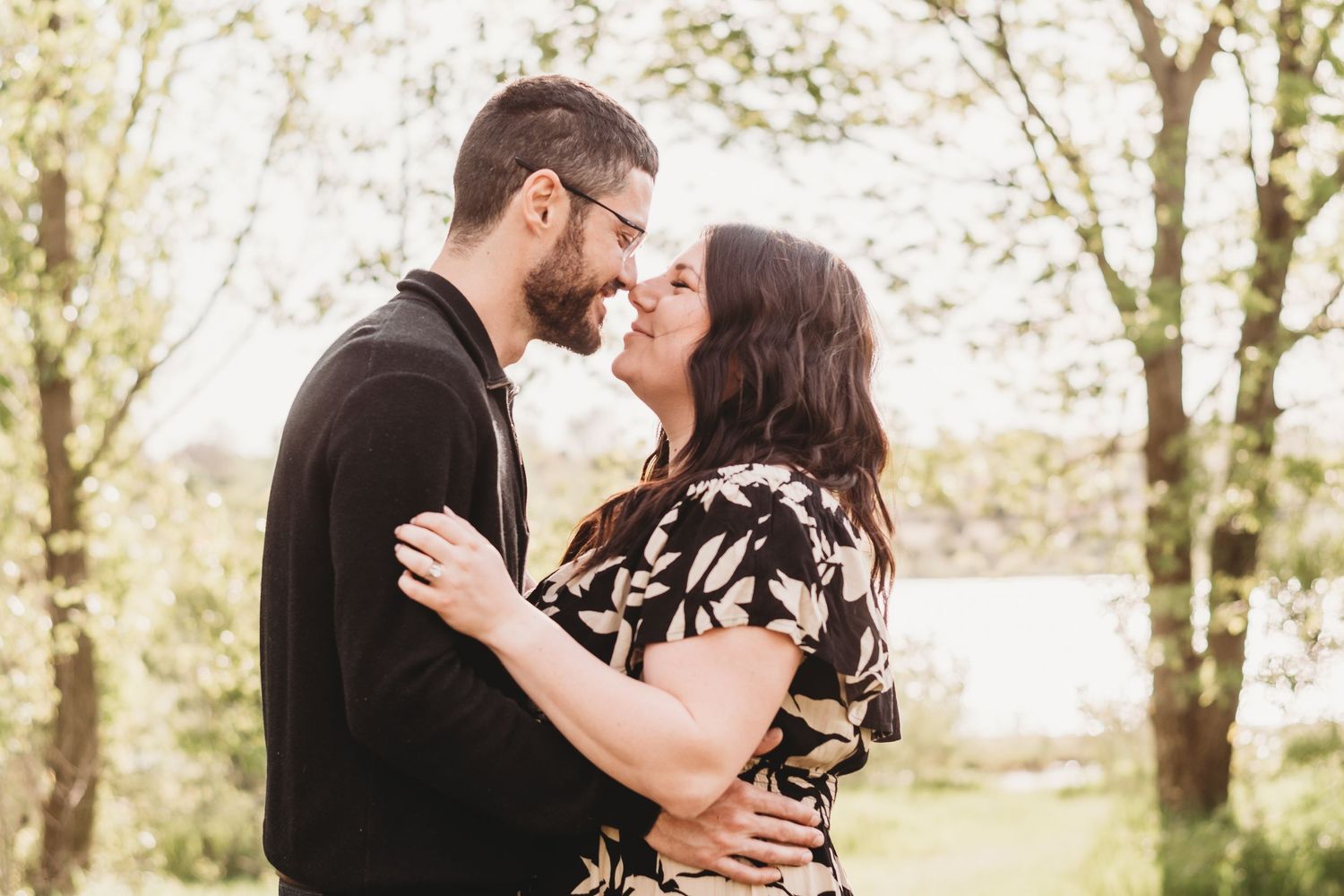 A romantic couple shares tender moments in a sunlit wooded area wearing black and floral attire.
