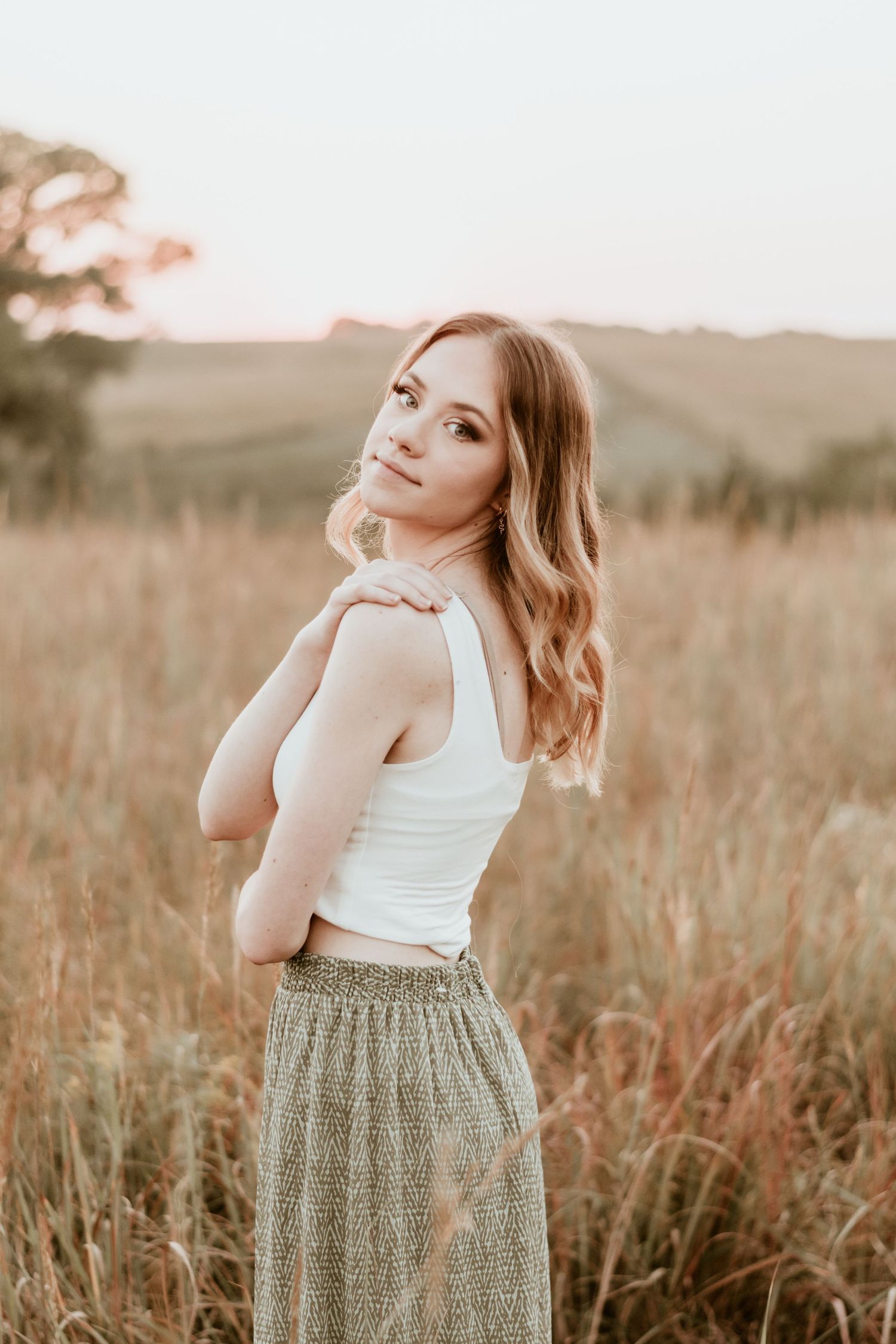 Person in white top and patterned skirt posing in golden field at sunset.