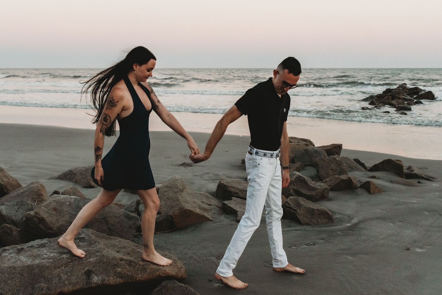 Couple holding hands walking barefoot on a rocky beach at sunset, woman in black dress, man in white jeans.