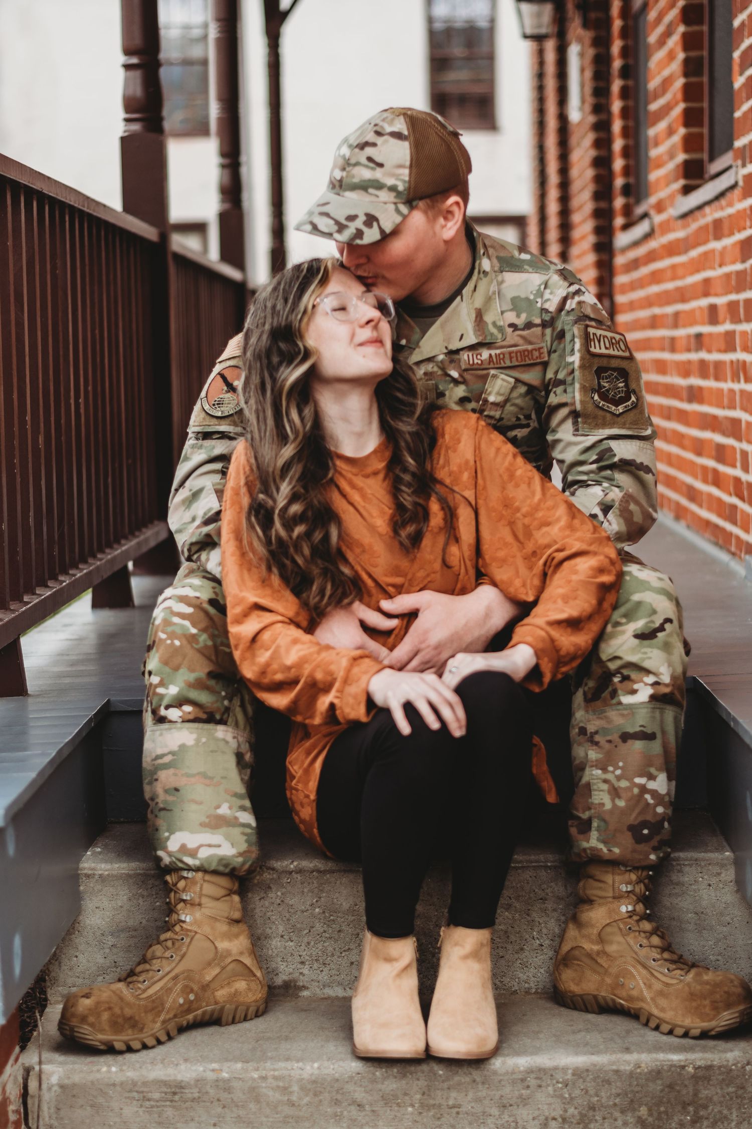 Military couple in camouflage uniform and orange sweater sitting together on brick steps sharing intimate moments.