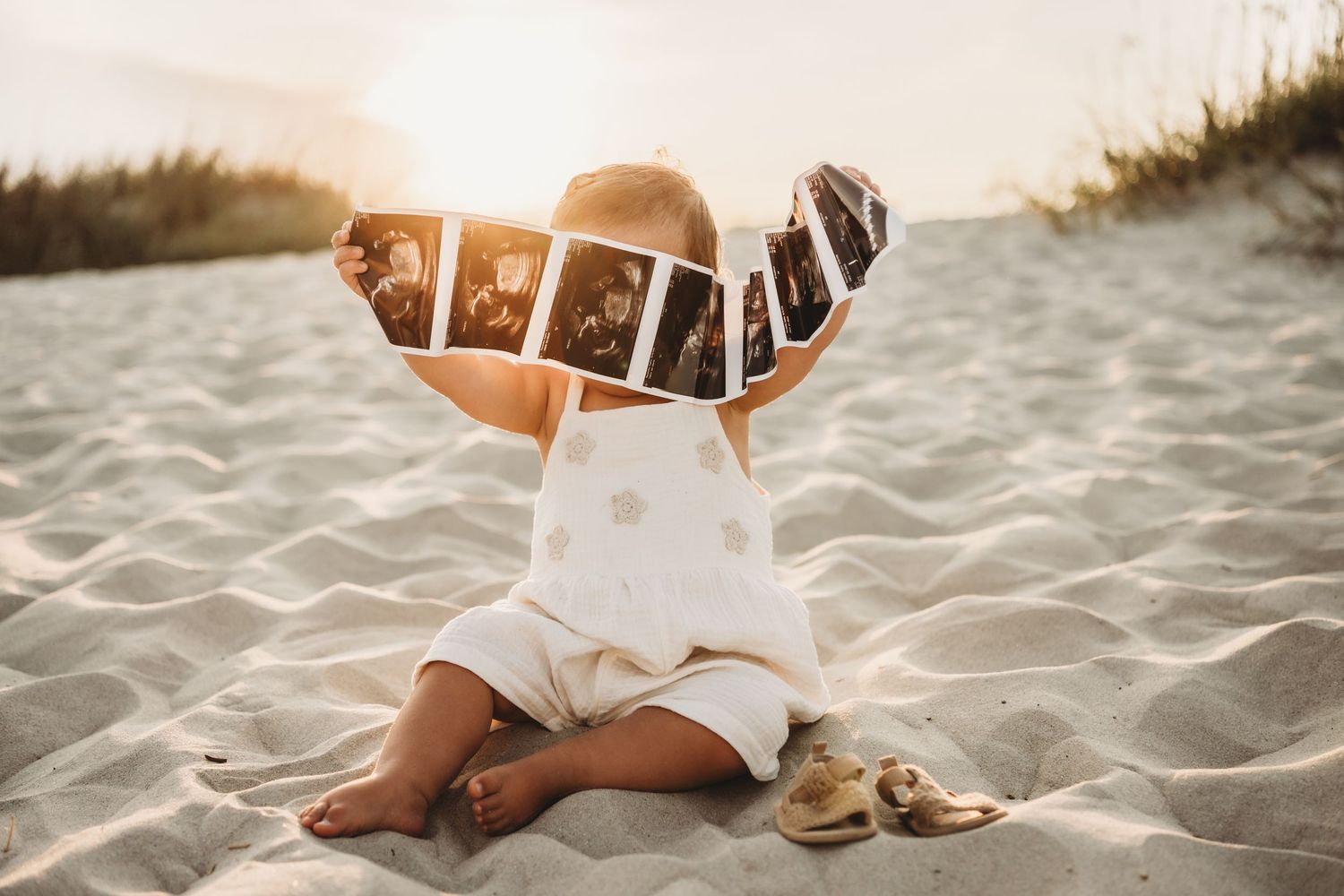 Baby sitting on sandy beach holding ultrasound photos in golden sunset light, wearing white polka dot outfit.