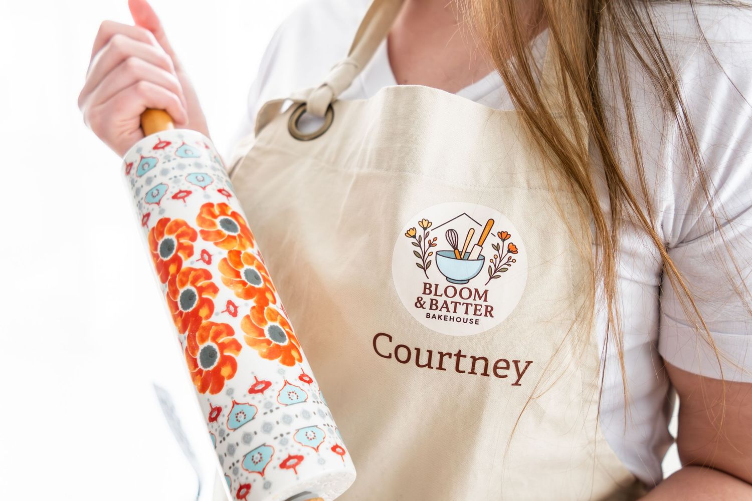Person wearing a personalized Bloom & Batter Bakehouse apron named Courtney, holding a floral rolling pin.