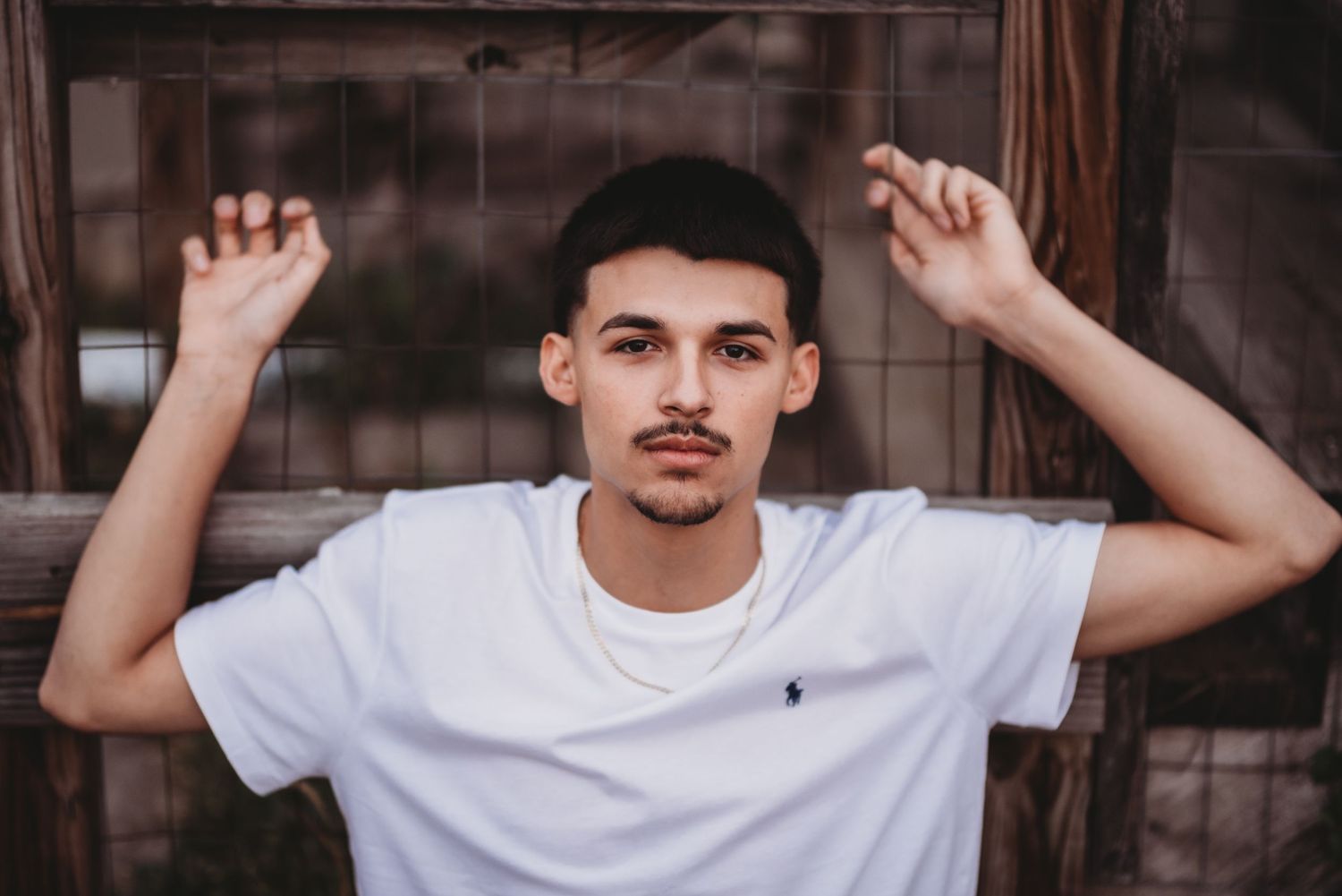 A person in a white t-shirt poses with raised arms against a dark textured wall.