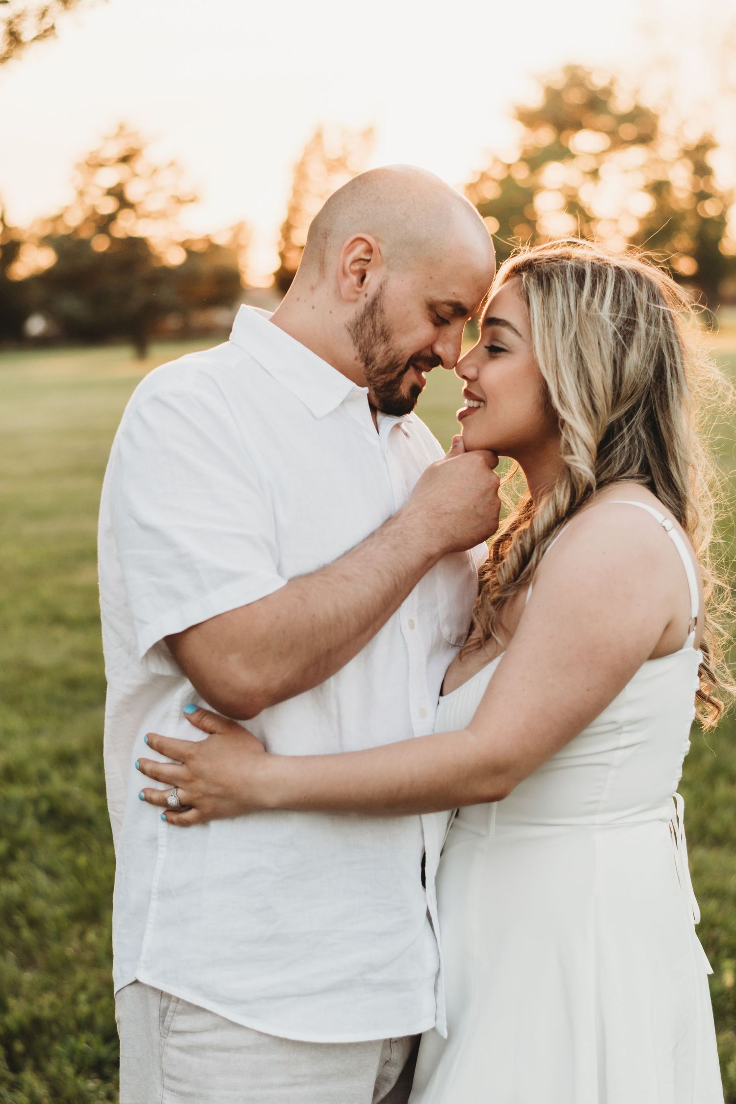 A couple in white clothing embraces during a golden sunset in a grassy field.