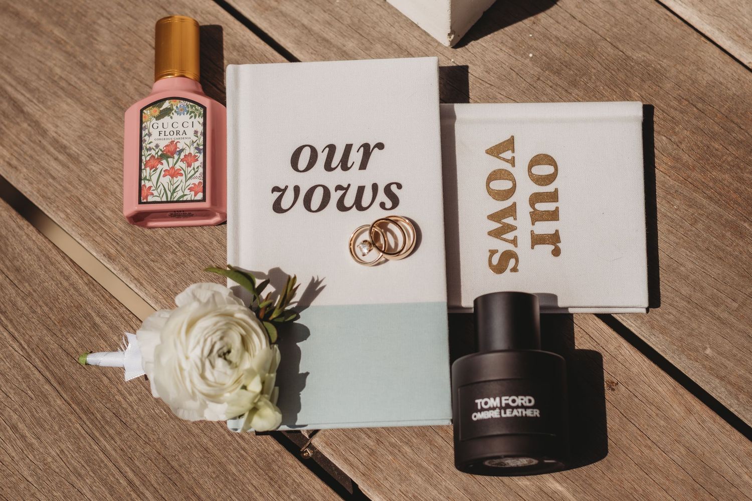 Wedding vow books, Gucci perfume, Tom Ford cologne, and a white flower arranged on a wooden surface.