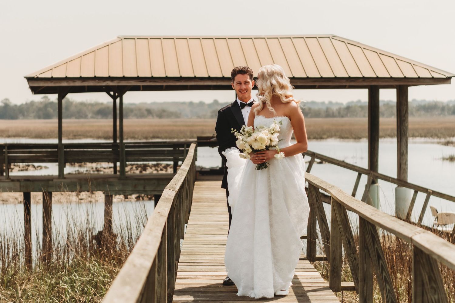 Bride and groom walking on wooden dock by waterfront gazebo on their wedding day.