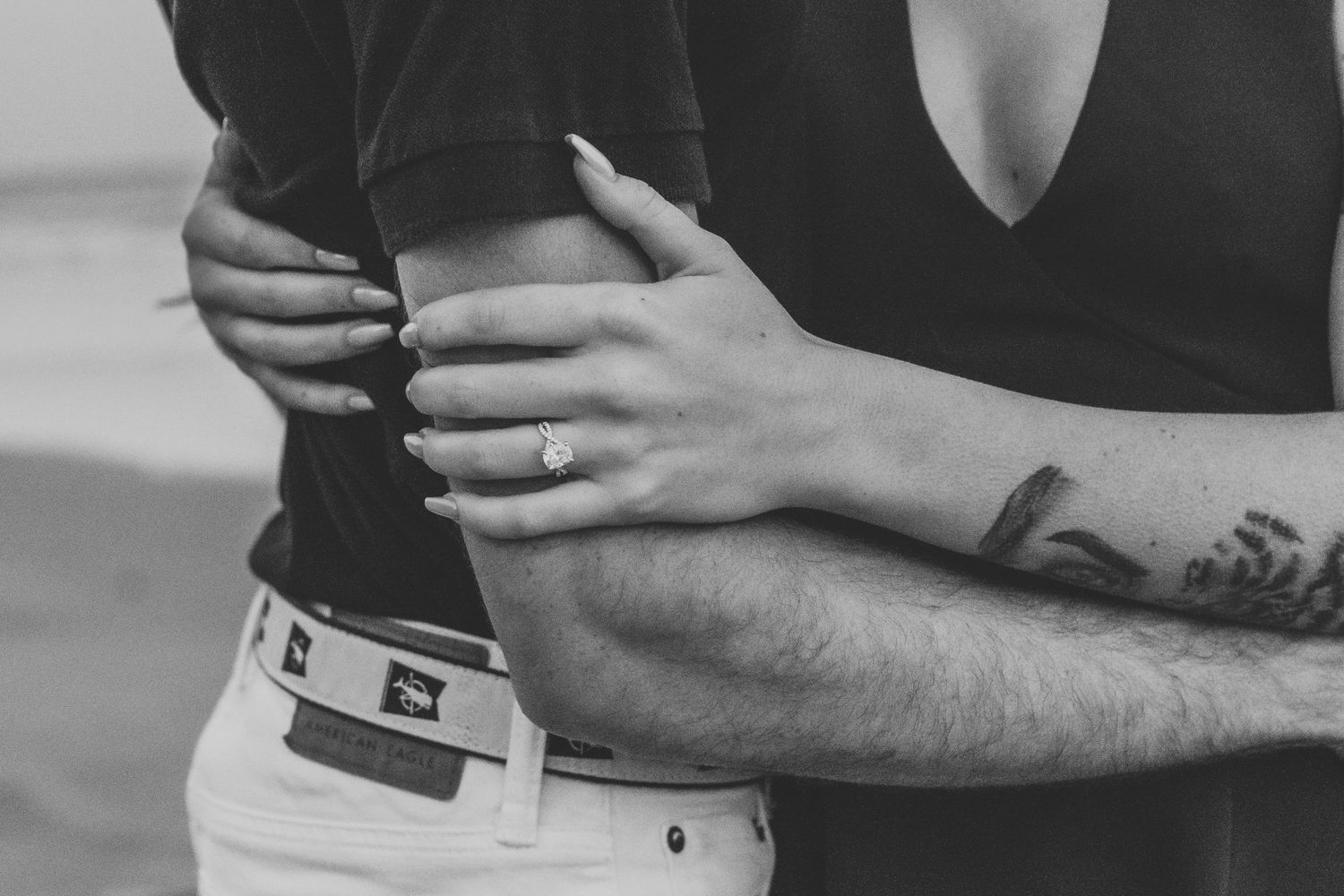 Black and white photo of a couple embracing, woman wearing engagement ring, man with tattoos, at the beach.