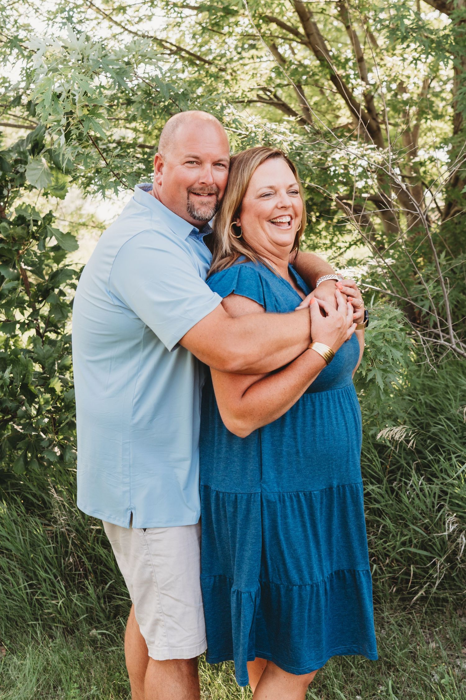 A couple embraces and laughs together in a sunny outdoor setting with green foliage.