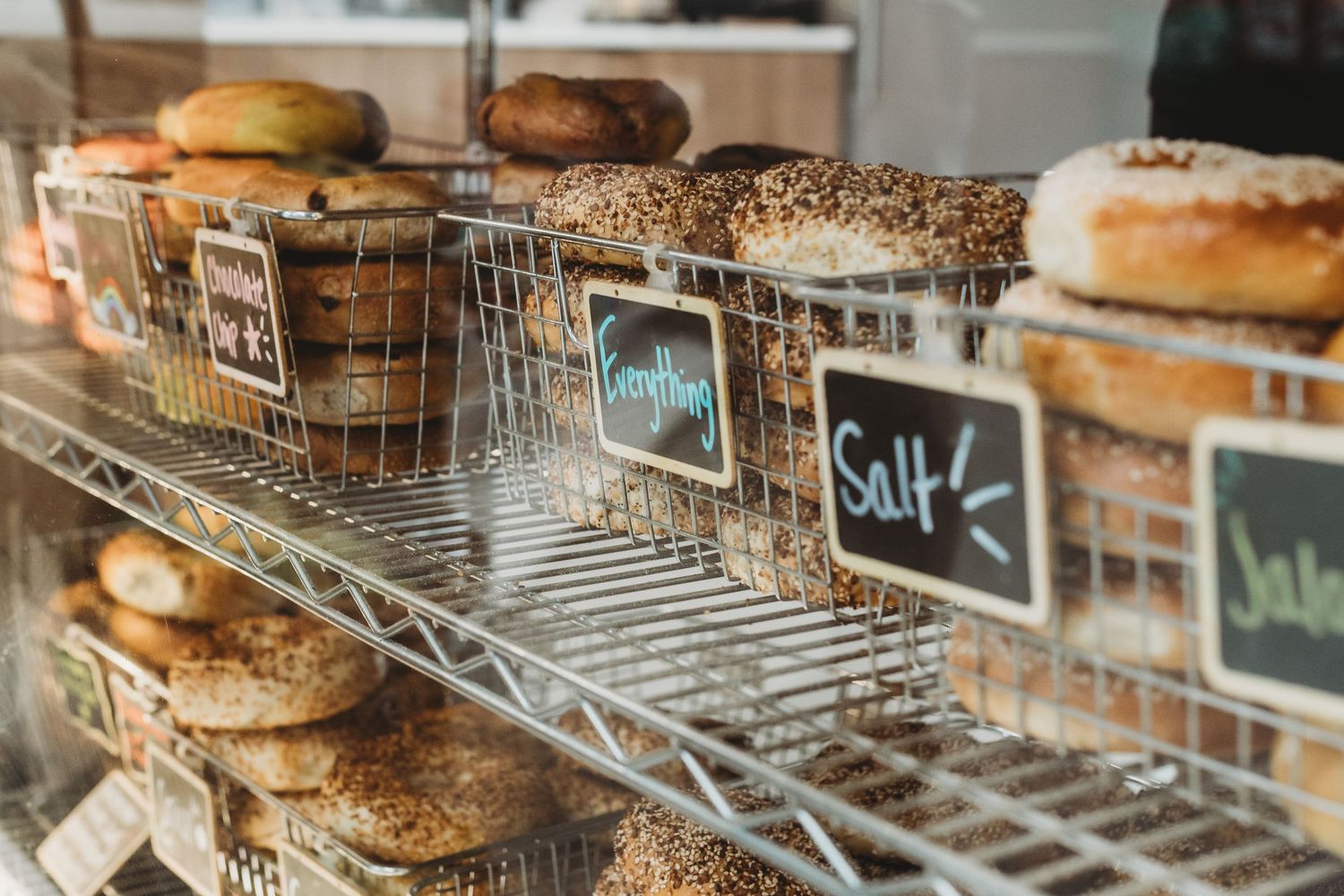 Fresh bagels displayed in wire baskets with chalkboard labels on metal shelving.