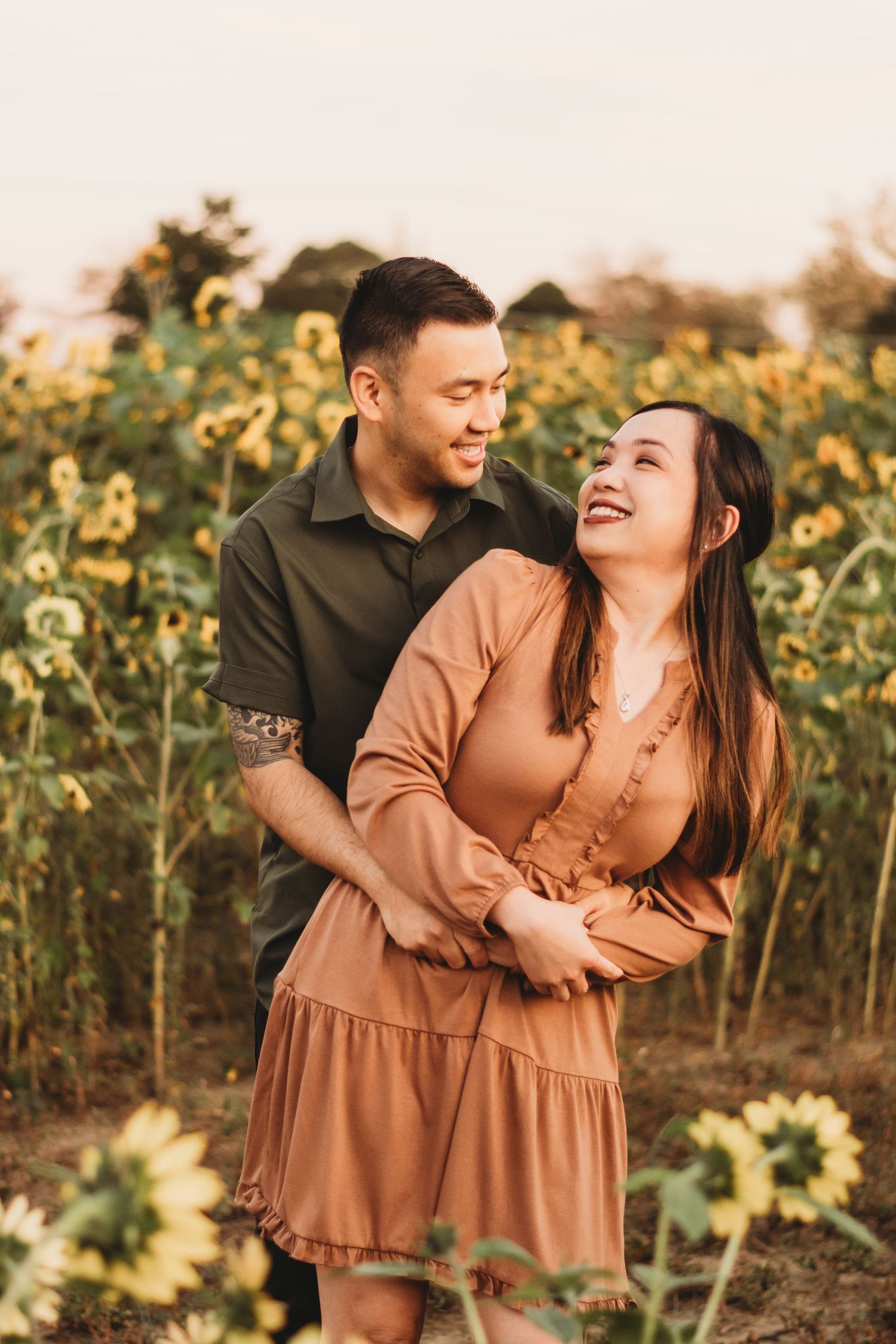 A couple laughs together in a sunflower field during golden hour.