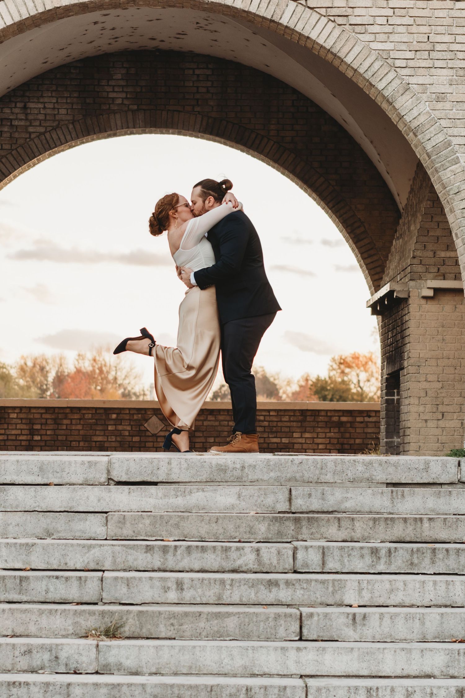 Couple sharing a romantic kiss on stone steps under a brick arch, woman in gold dress lifting her heel.