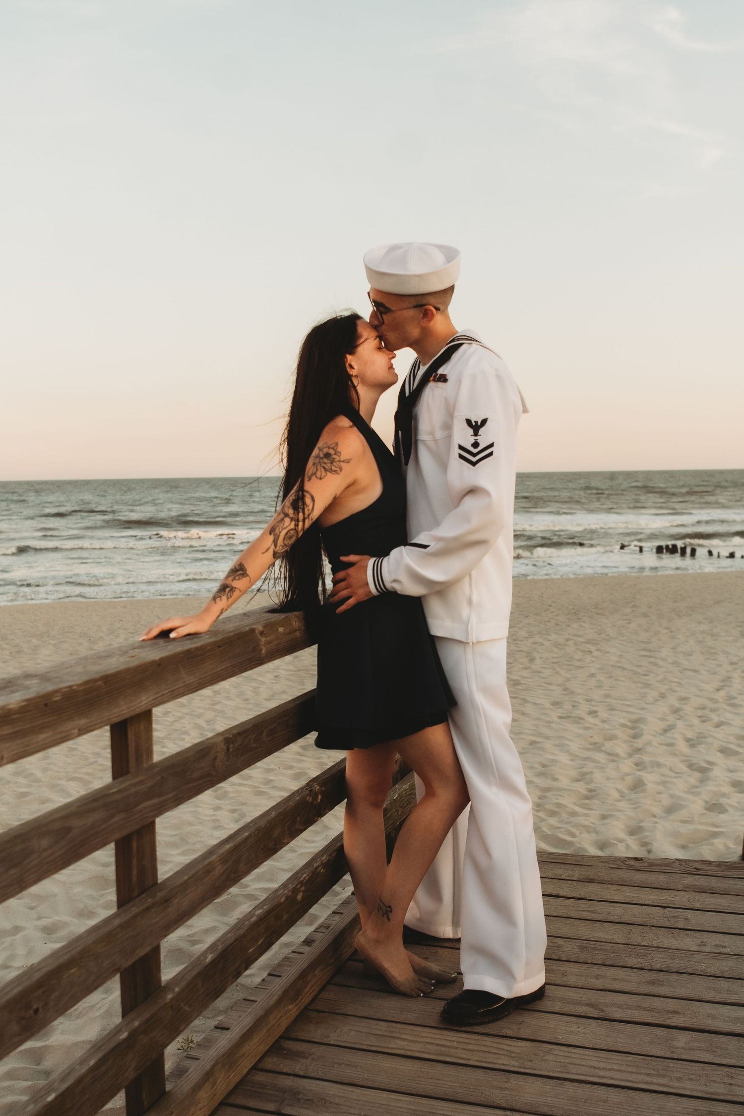 A sailor in white uniform kisses his partner on a wooden boardwalk by the beach at sunset.