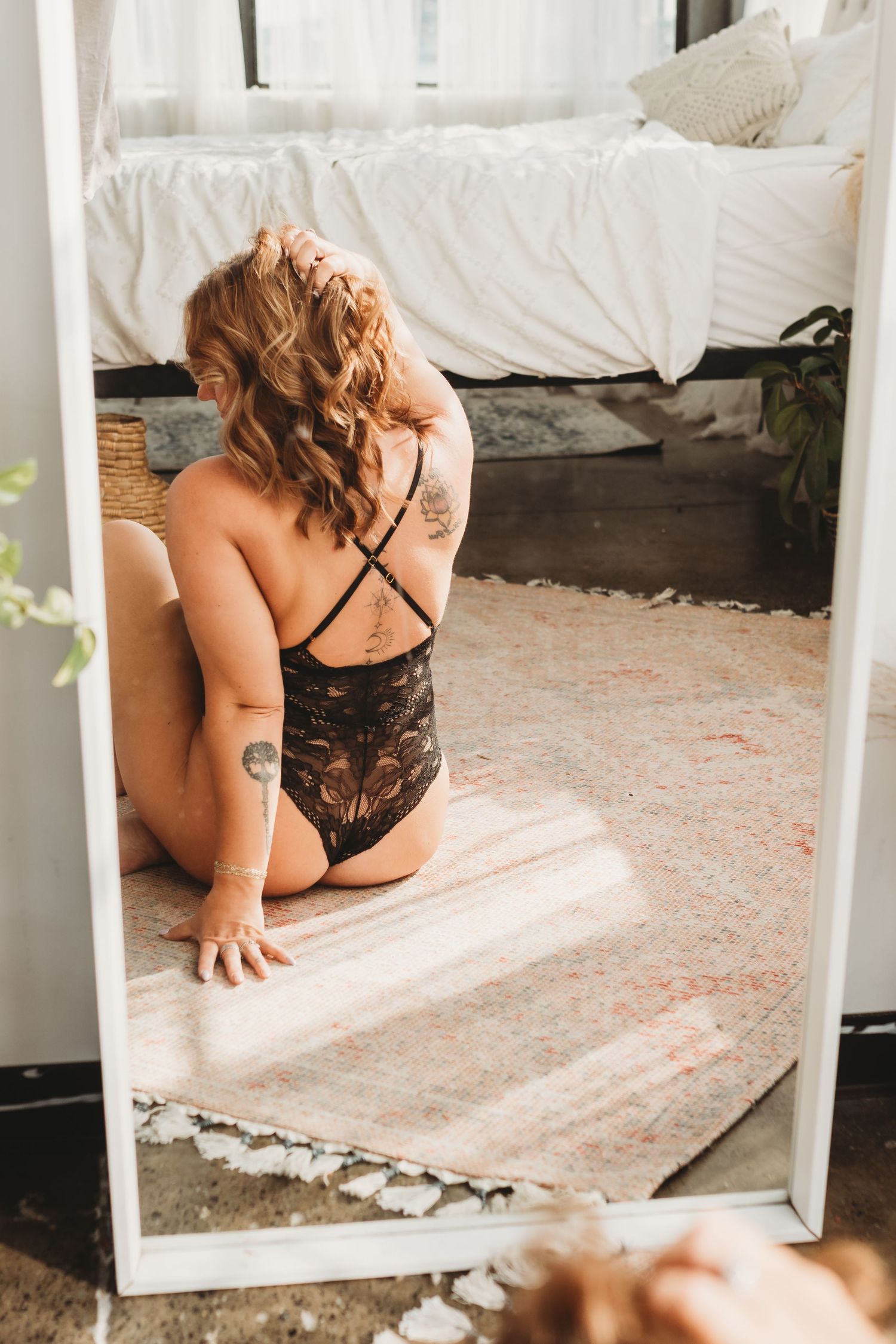 Woman with tattoos in black lace bodysuit sitting on rug reflected in large floor mirror in boho bedroom.