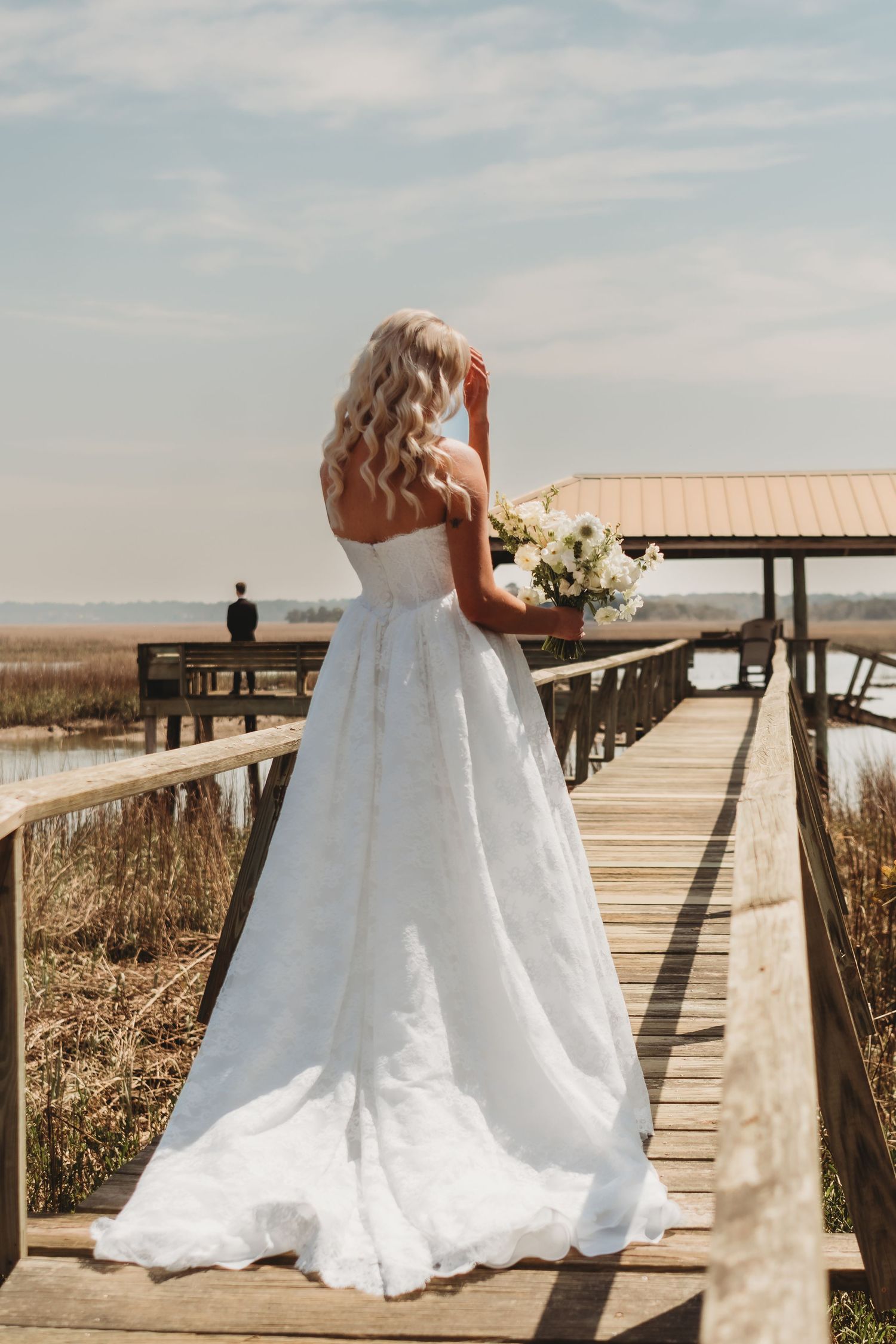 Blonde bride in white ball gown holds bouquet on wooden boardwalk over marsh, back to camera, on her wedding day.