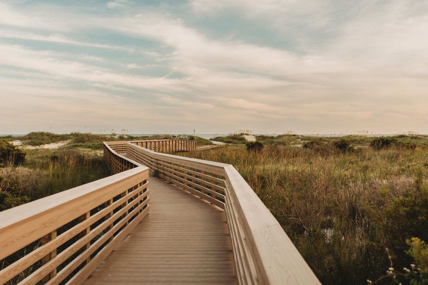 Wooden boardwalk path extends through coastal dunes at sunset.