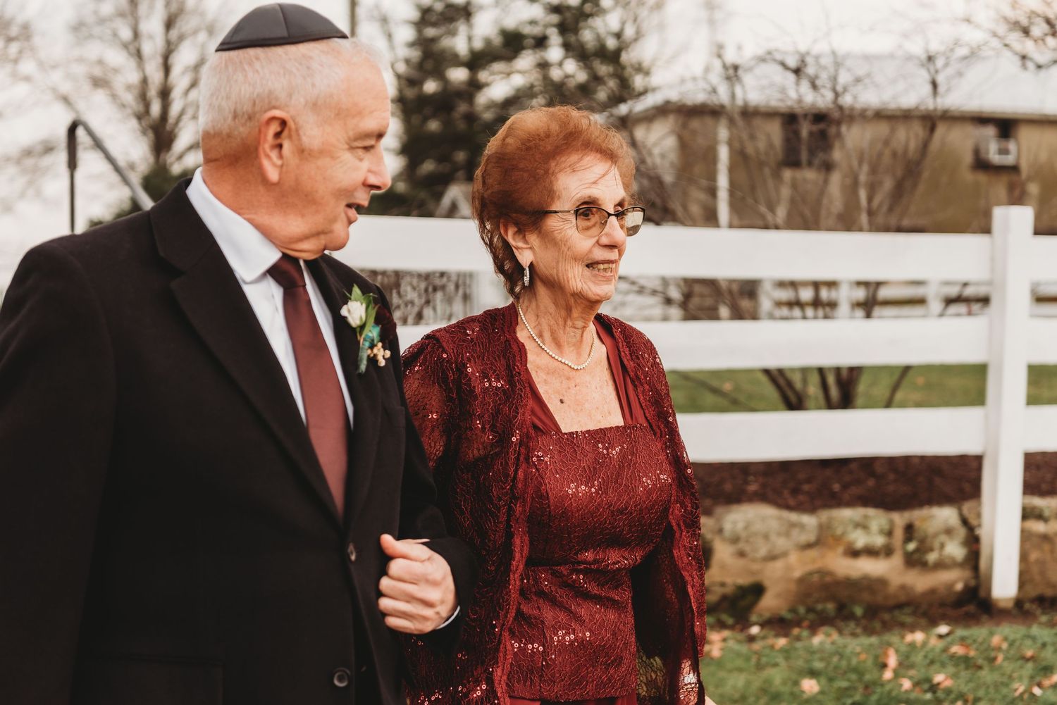 Couple in formal attire sharing joyful moments outdoors near white fence during autumn.