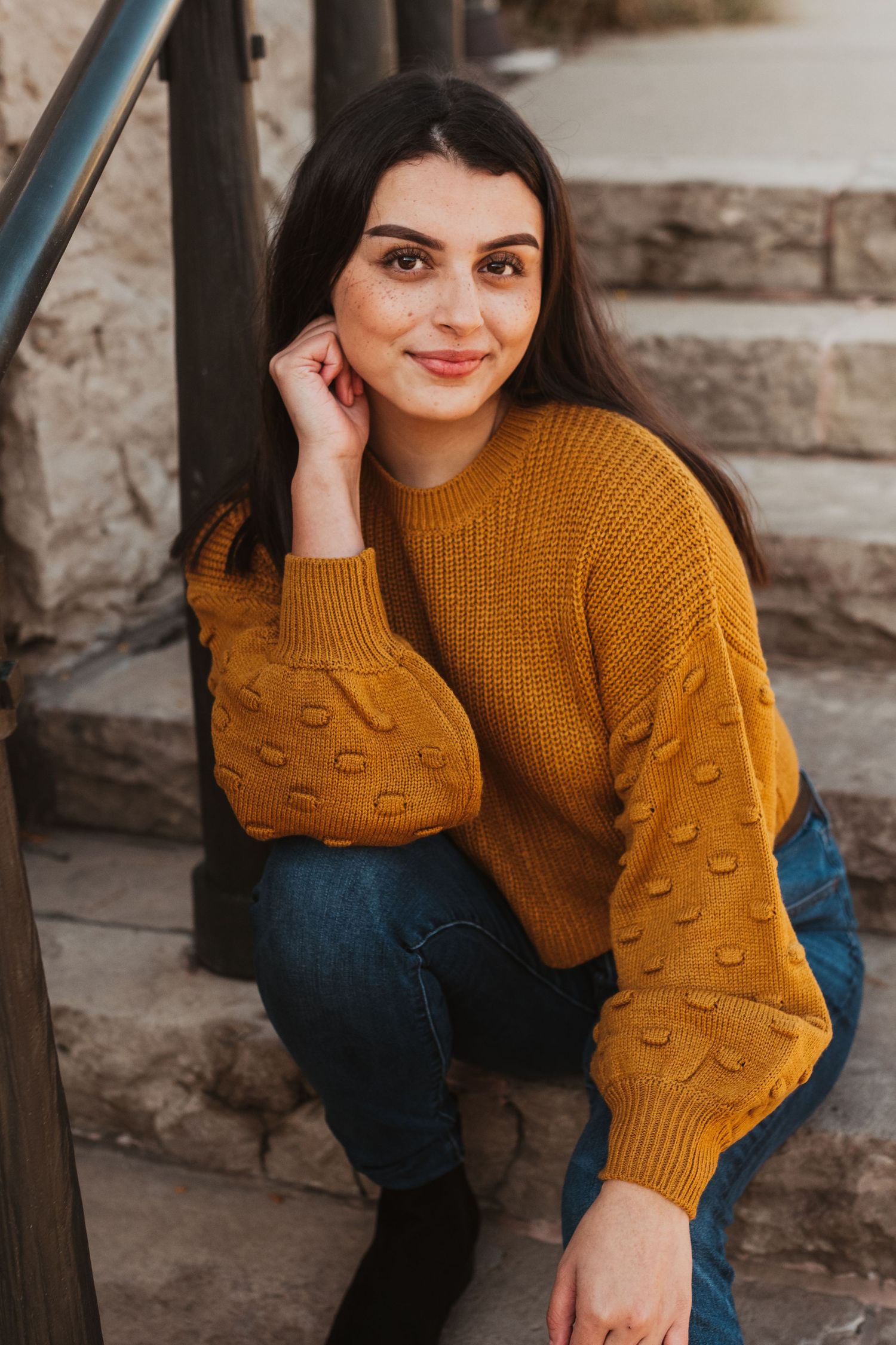 A person wearing a cozy mustard yellow knit sweater with textured sleeves sits casually against a stone wall.
