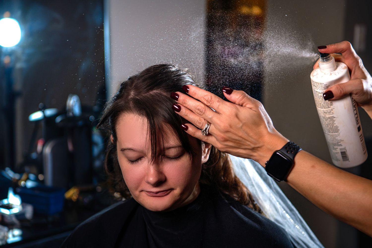 Hairstylist applying hairspray to a brunette bride during wedding day preparation in a salon.