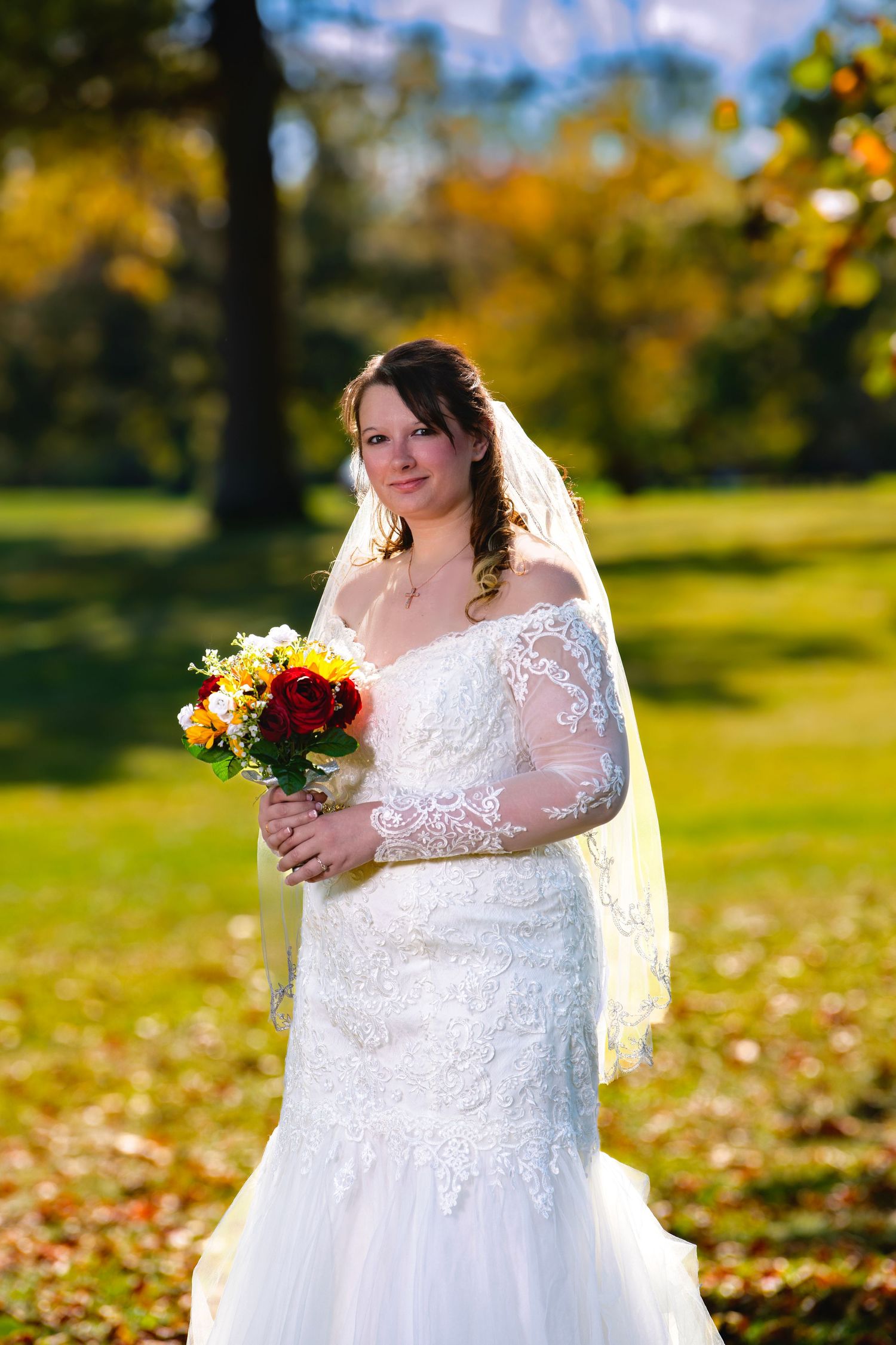 Beautiful bride in a lace mermaid wedding gown holding a vibrant bouquet of red and yellow flowers in an autumn park setting.