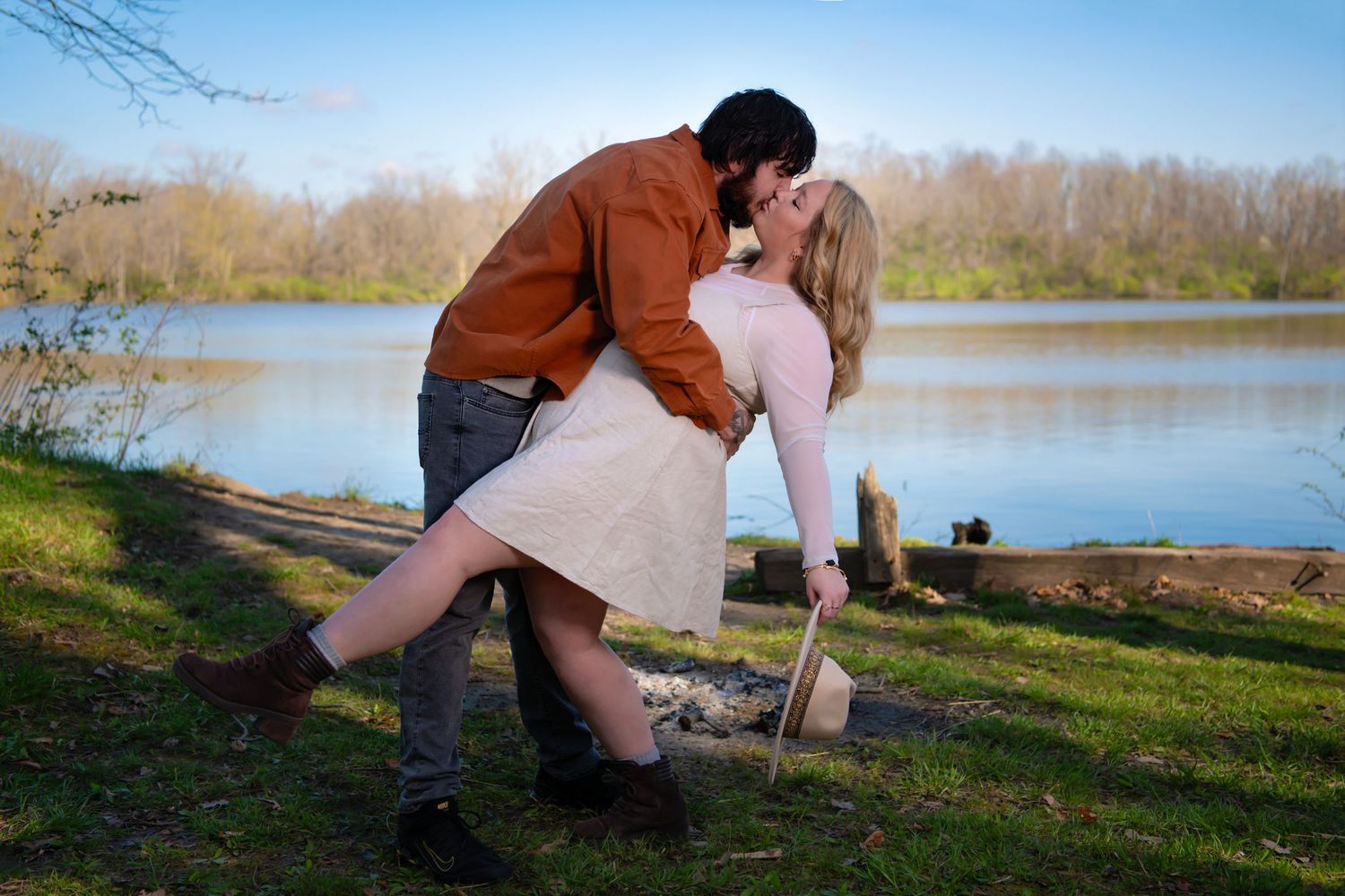 Couple sharing a romantic dip kiss by a serene lake surrounded by lush green trees.