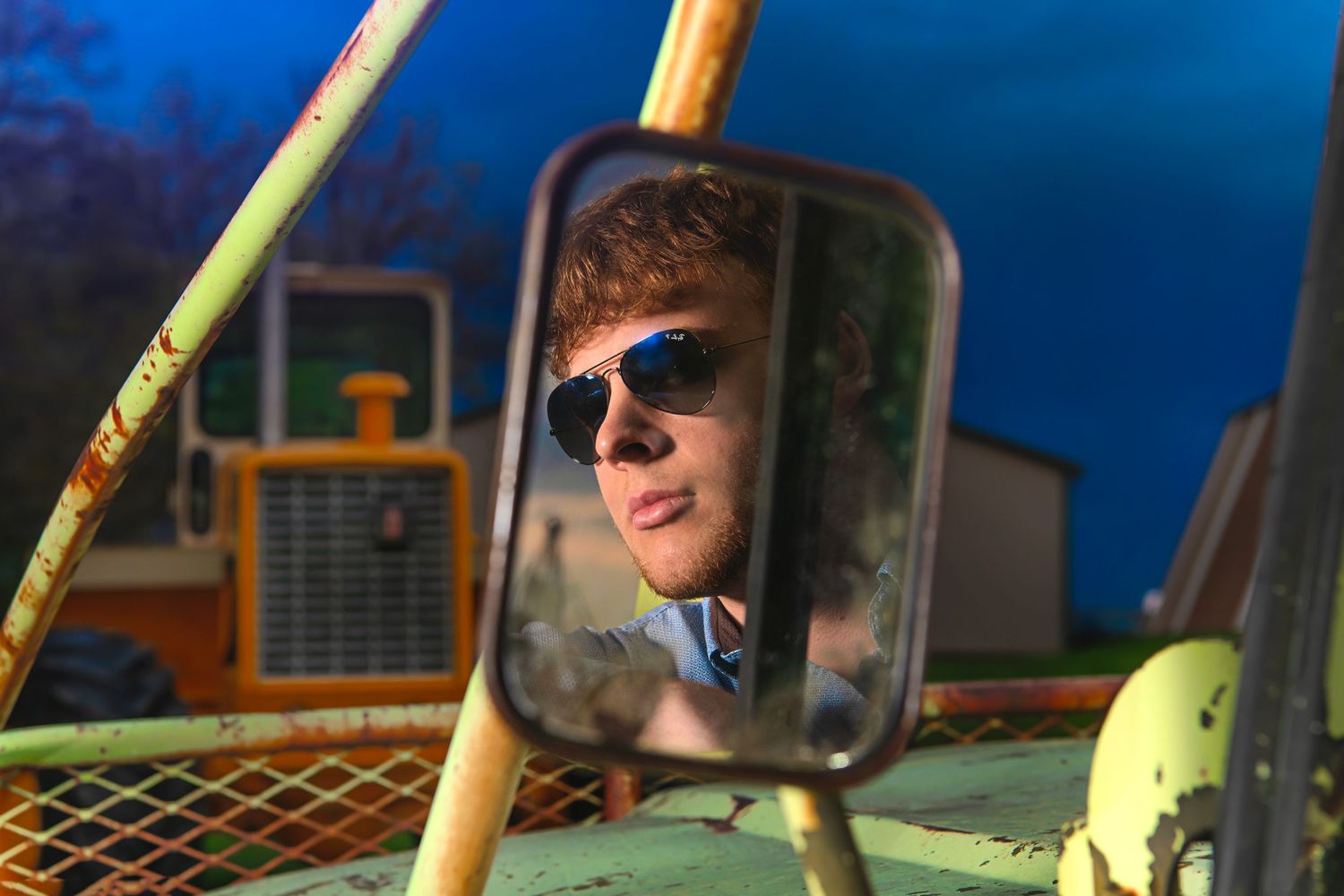 Young man with sunglasses reflected in vintage green tractor side mirror at dusk.