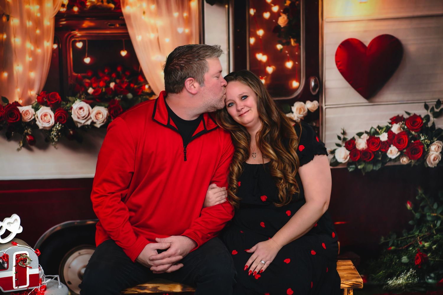 Couple sharing a romantic kiss during a Valentine's Day photo session with red roses, hearts, and warm string lights backdrop.