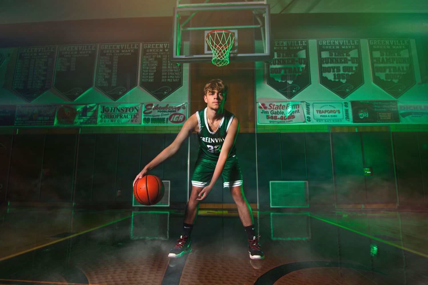 Young athlete in a green basketball uniform poses confidently with a ball in a dramatic smoke-filled school gymnasium.