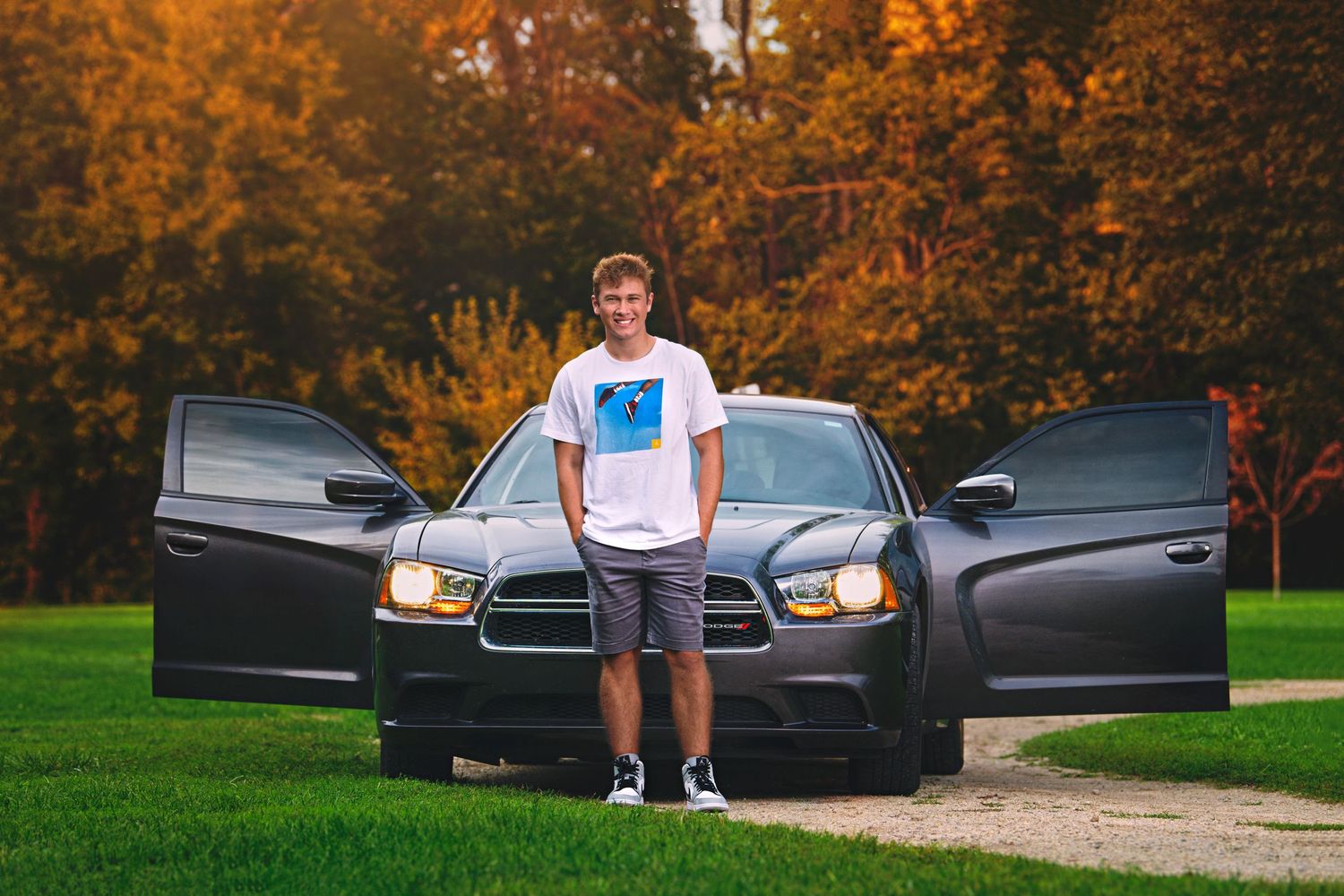 Teen boy in white tee stands between open doors of black Dodge Charger amid autumn foliage.