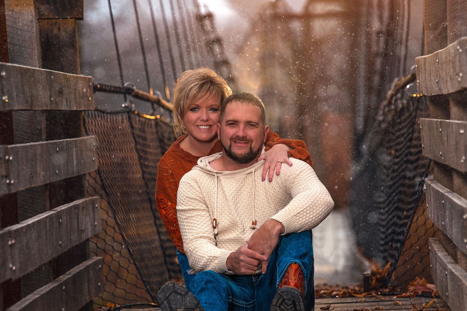 Smiling couple sitting together on a rustic rope bridge surrounded by autumn leaves and mist.