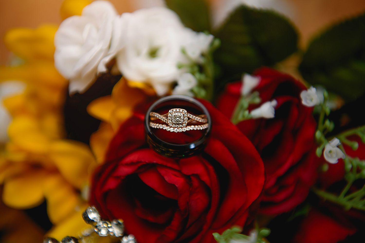 Close-up of elegant gold engagement and wedding rings displayed on a red rose in a bridal bouquet with colorful flowers.