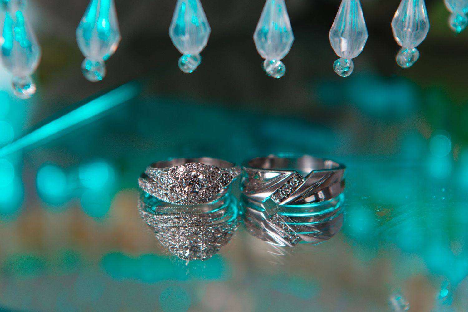 Two silver diamond wedding rings displayed on a reflective teal surface with crystal chandelier drops above.