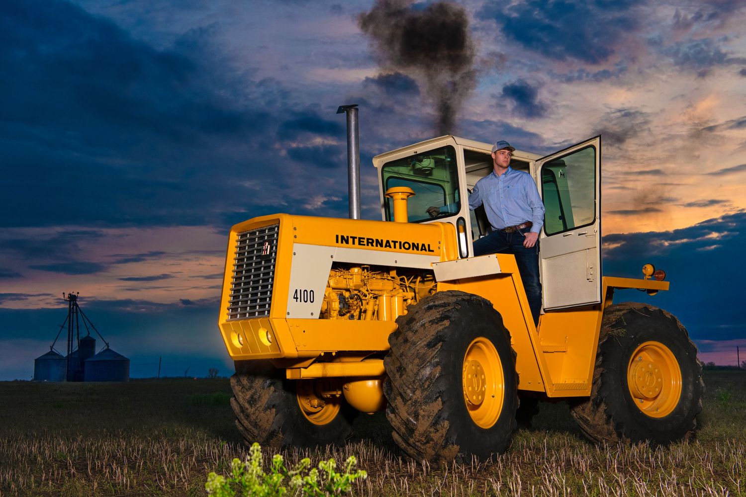 Man standing on yellow International 4WD tractor in field under dramatic stormy sunset sky.