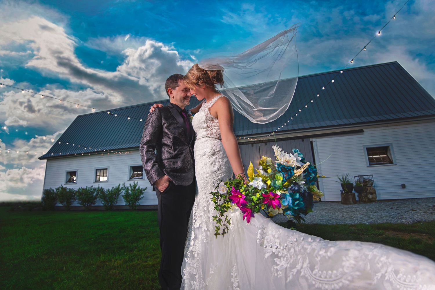 Bride and groom sharing a tender kiss outdoors near a white barn under dramatic cloudy skies.