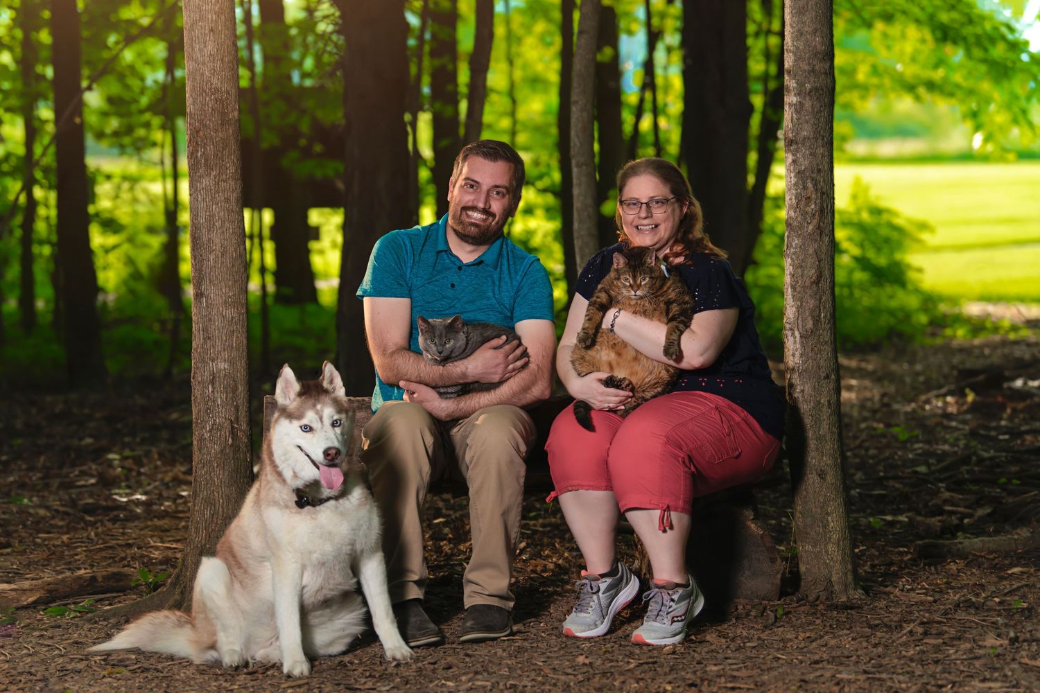 Couple sitting in forest with Husky dog and two cats in dappled sunlight.