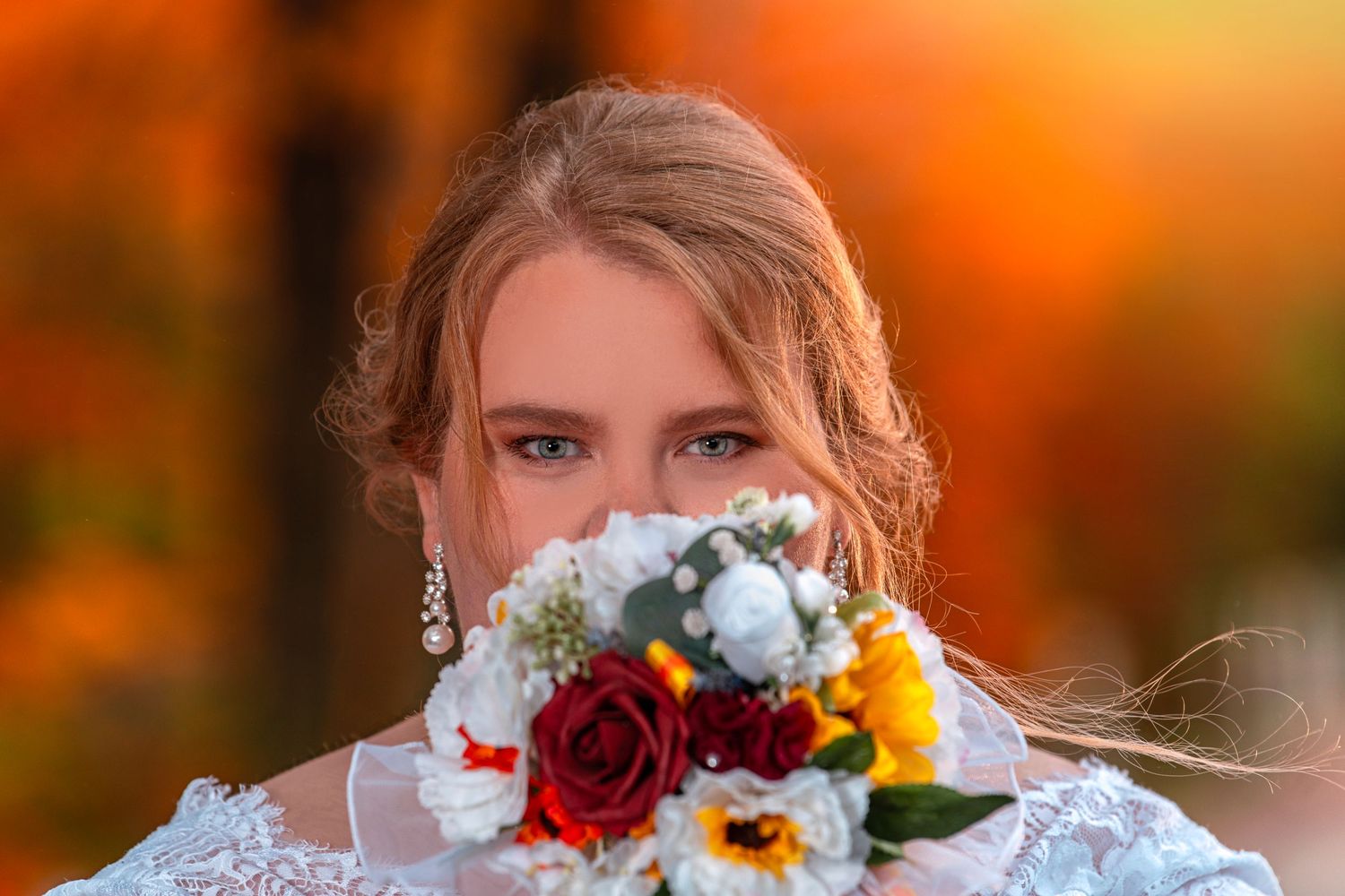 Bride holding a colorful bouquet of red roses and white flowers in front of her face with autumn foliage background.
