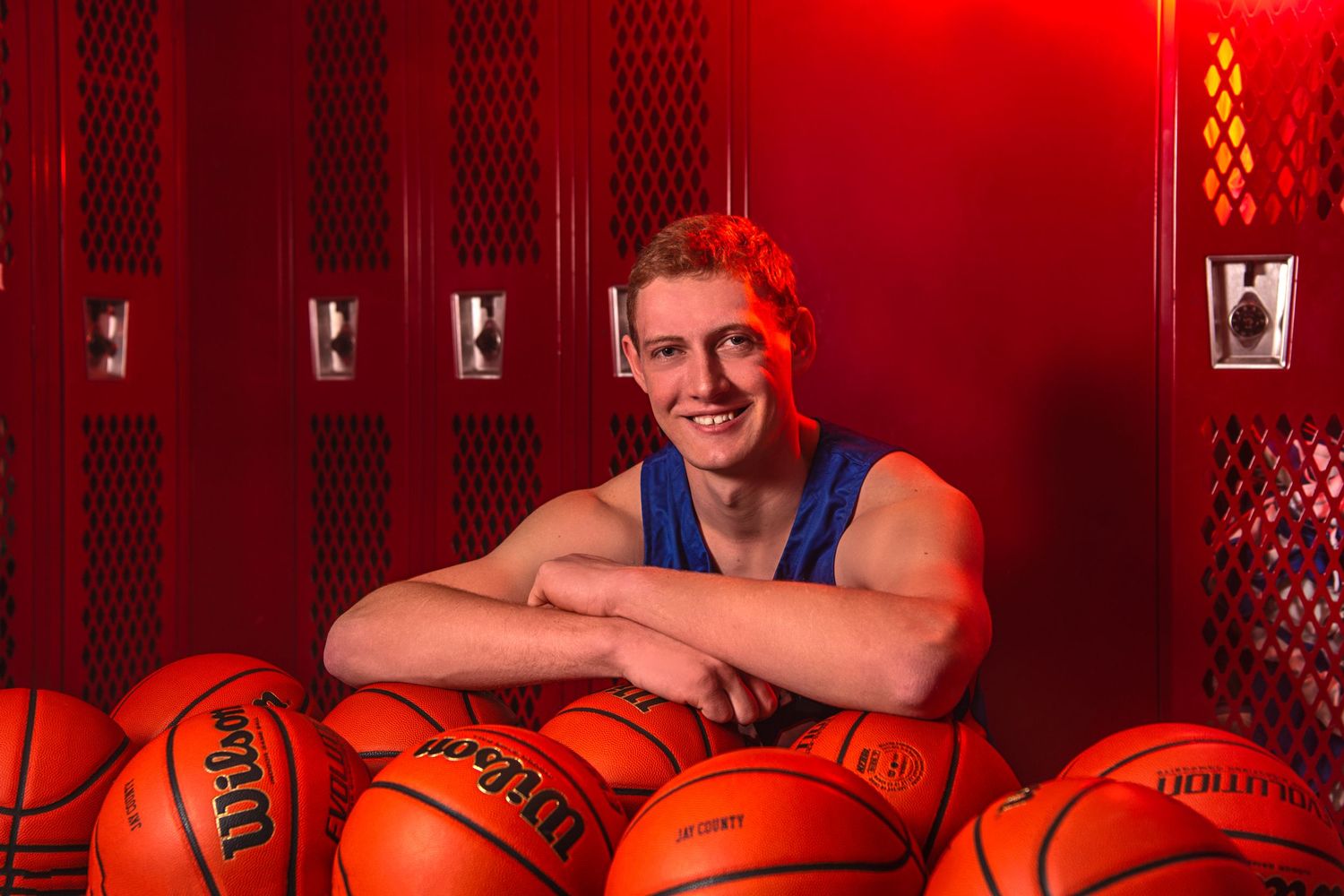 Smiling young male basketball player posing with Wilson basketballs in red-lit locker room.