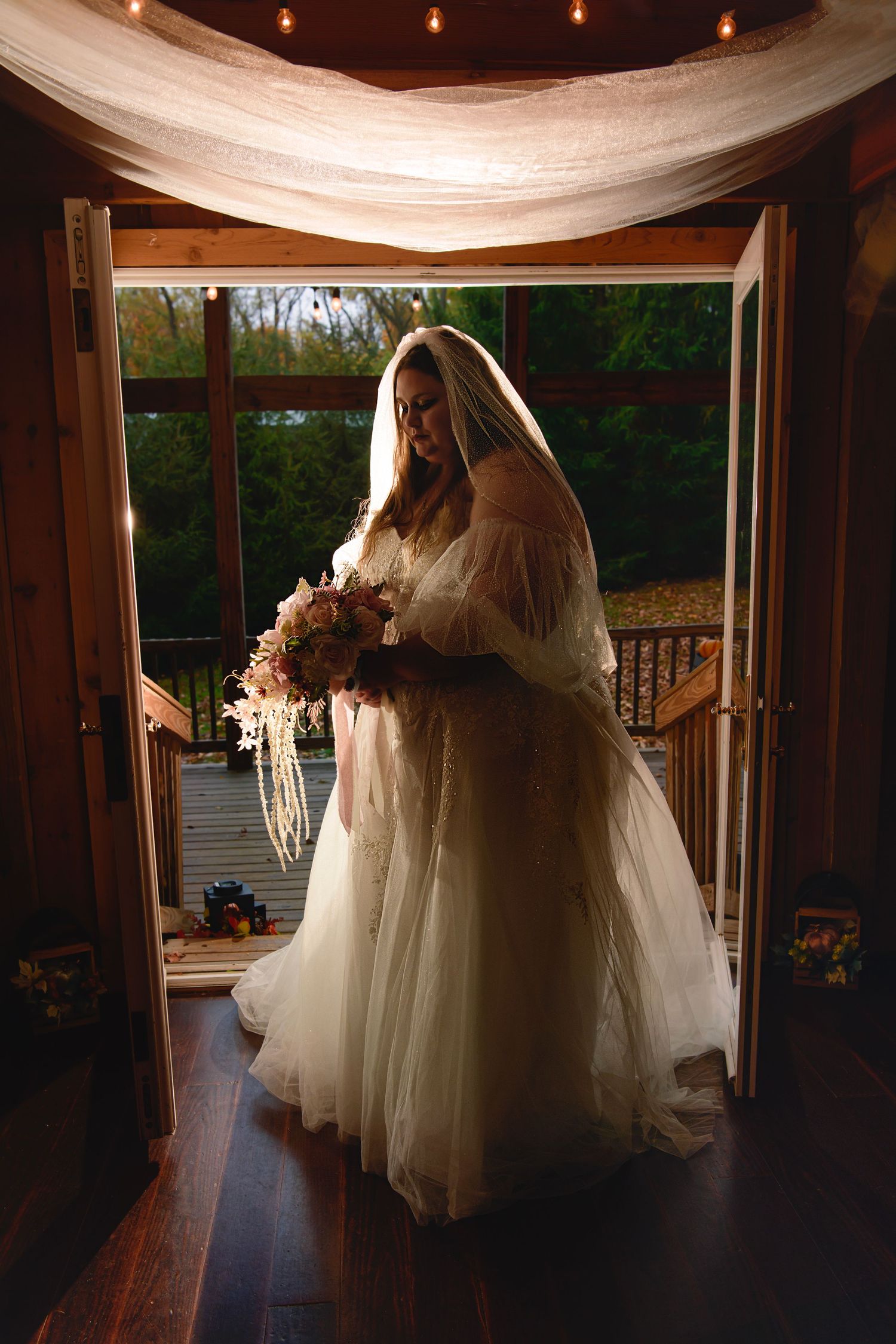 Bride in a flowing white tulle gown and veil holding a bouquet, standing in an open doorway at dusk.