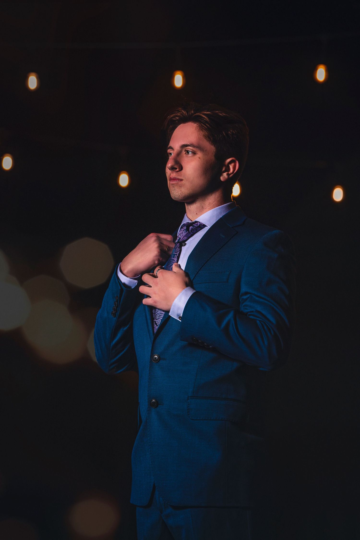 Stylish man in blue suit adjusting tie with bokeh string lights in dark background.