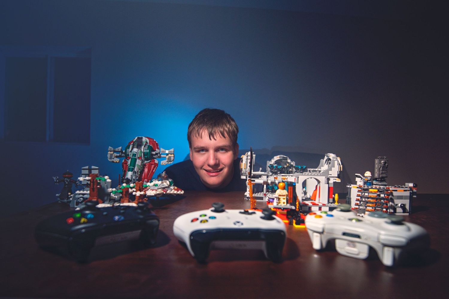 Man smiling behind a table filled with LEGO sets and gaming controllers in blue studio lighting.
