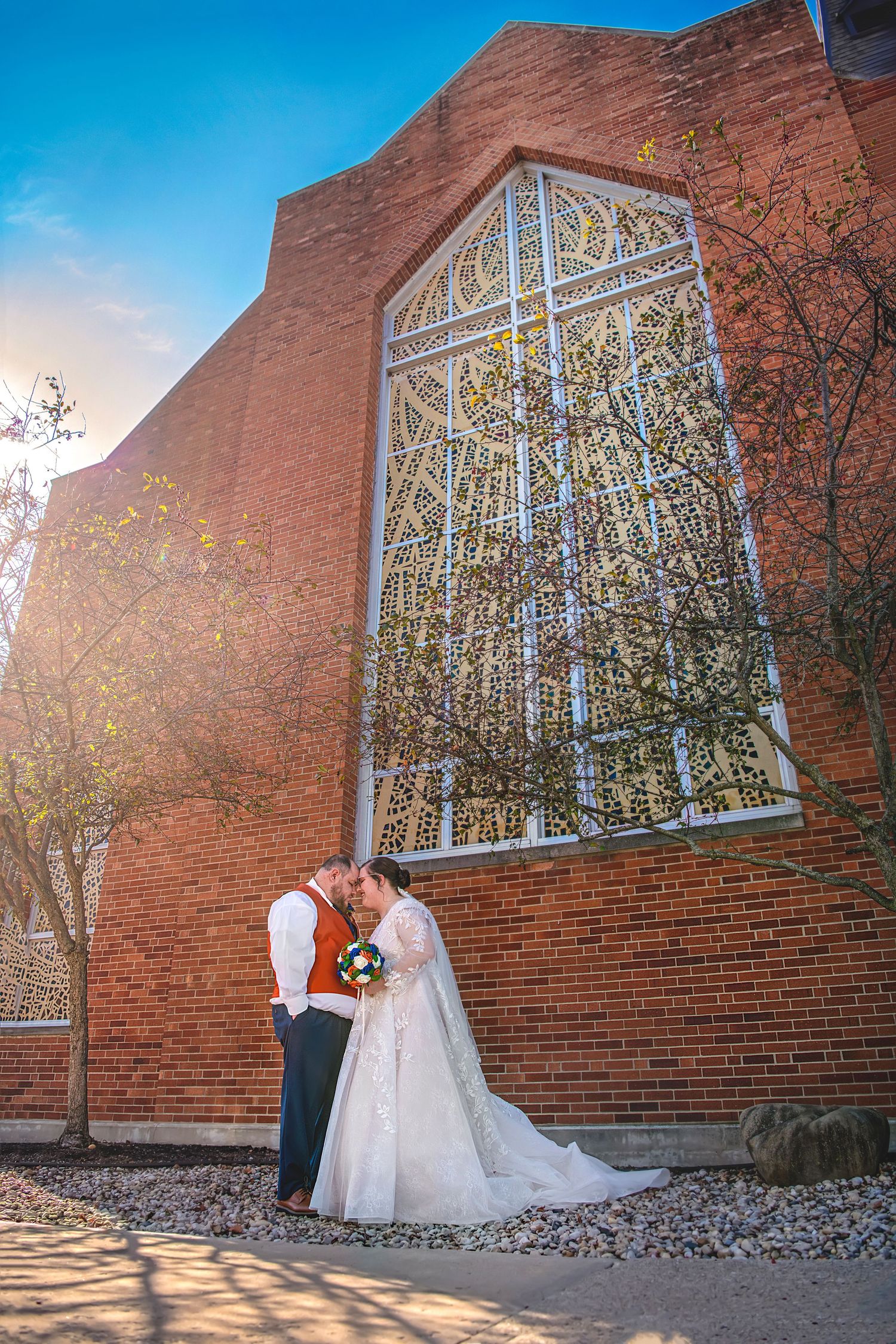 Bride and groom kissing outside brick church with ornate window and golden sunset light.